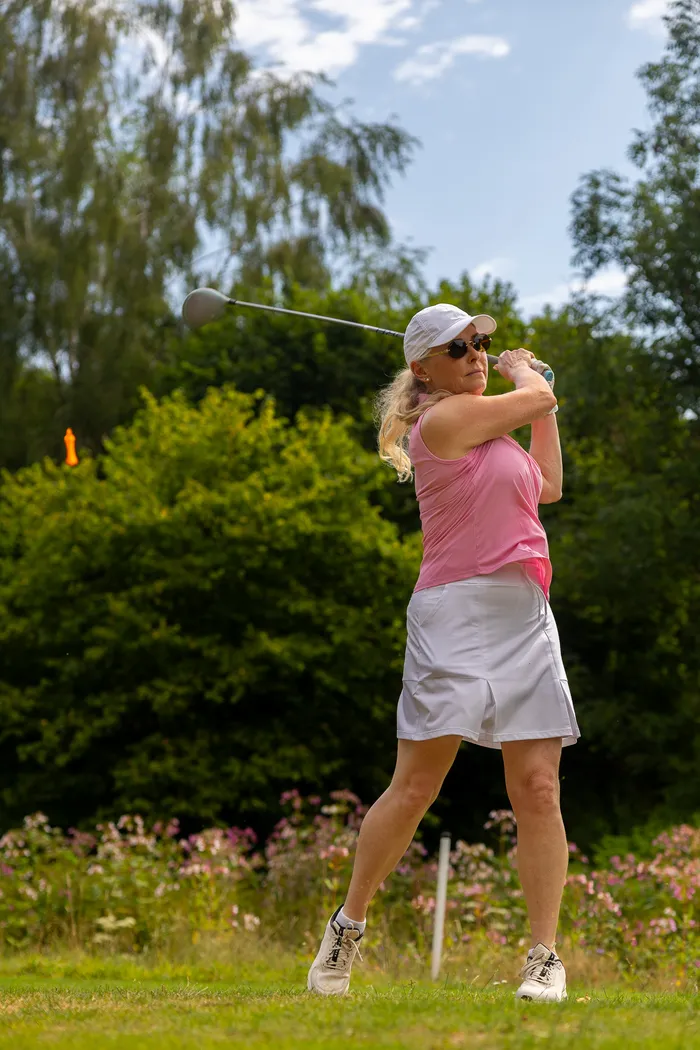 Eine Frau in rosa Oberteil und weißem Rock nach einem Golfschwung auf einem Platz mit Bäumen im Hintergrund.
