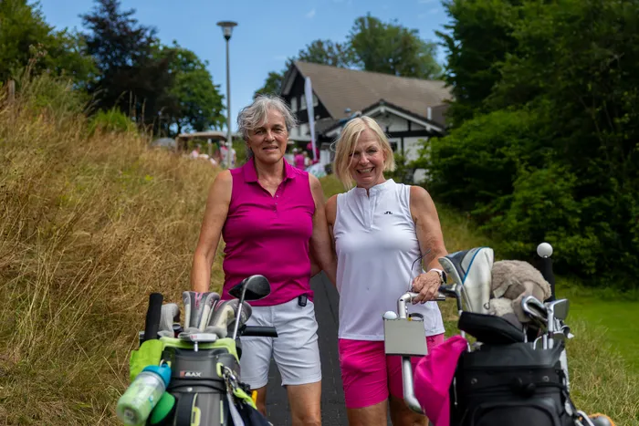 Zwei lächelnde Golferinnen stehen mit ihren Golfbags auf einem Weg.