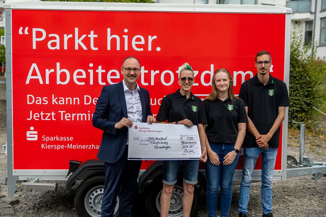 Vier Personen halten gemeinsam einen großen Scheck vor einem roten Anhänger der Sparkasse Kierspe-Meinerzhagen.