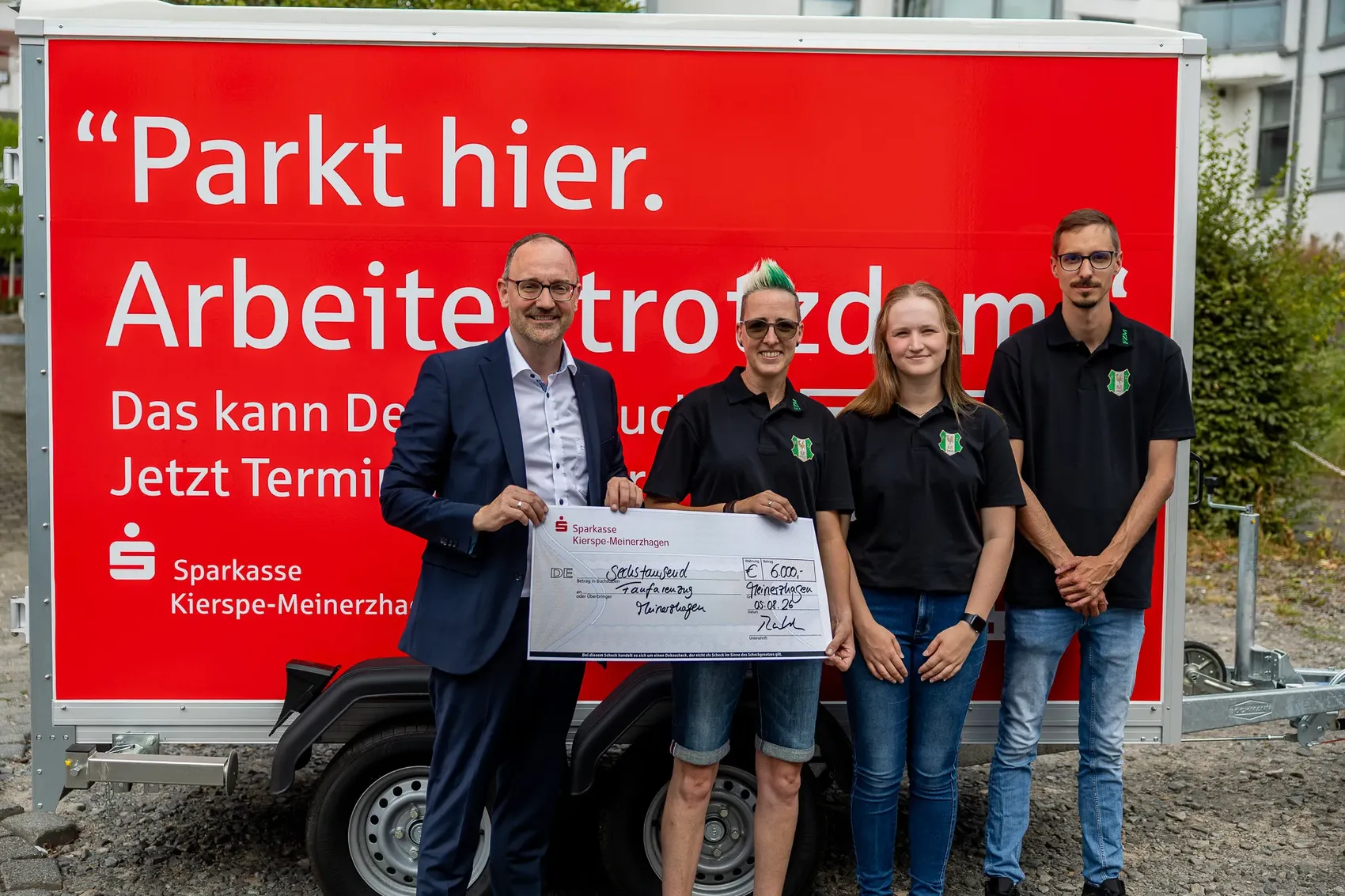 Vier Personen halten gemeinsam einen großen Scheck vor einem roten Anhänger der Sparkasse Kierspe-Meinerzhagen.