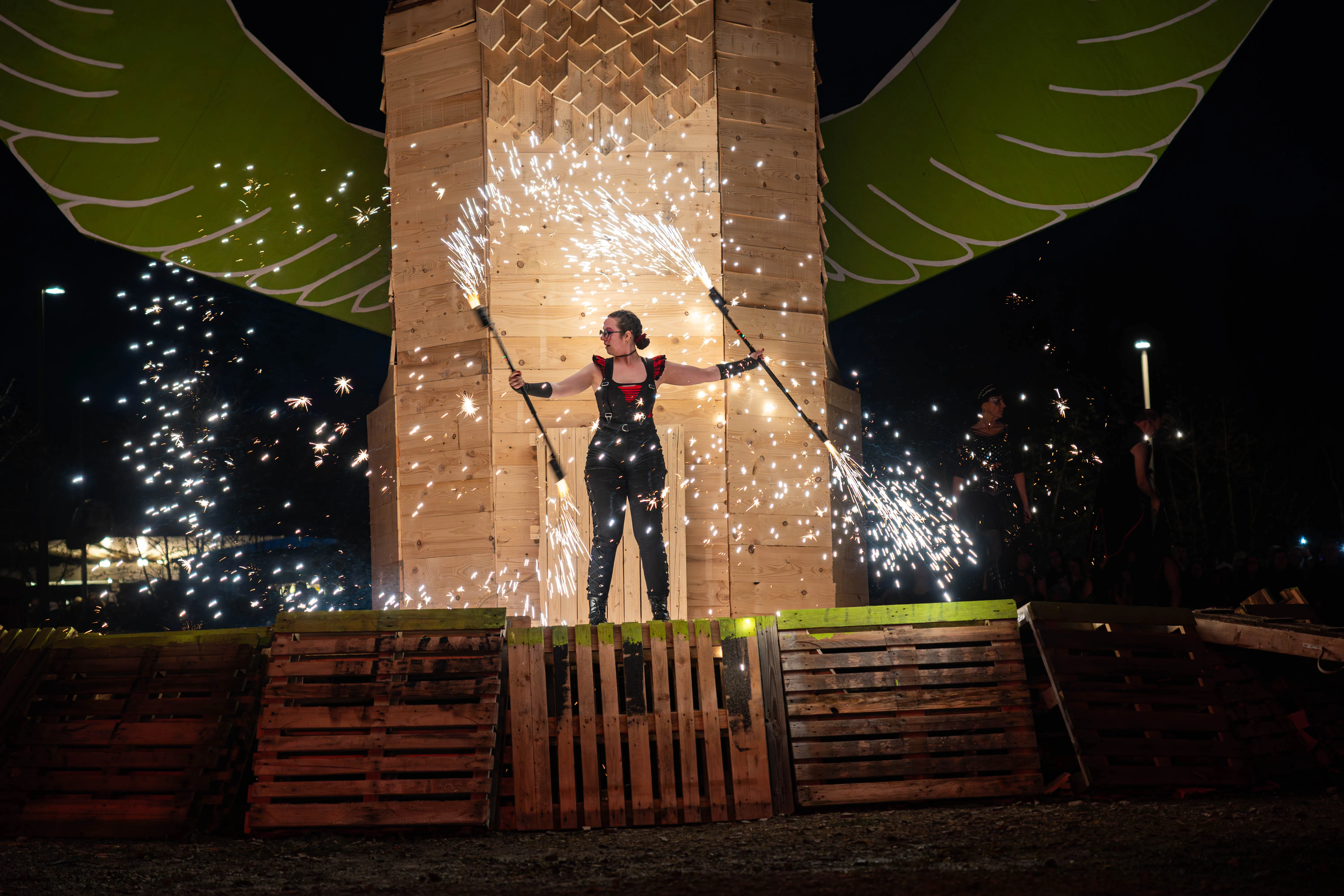 Eine Frau mit rotem Kopftuch und schwarzer Kleidung steht auf einer Palettenkonstruktion und schwingt zwei brennende Stäbe, die Funken sprühen. Im Hintergrund eine Holzkonstruktion mit angedeuteten Flügeln.