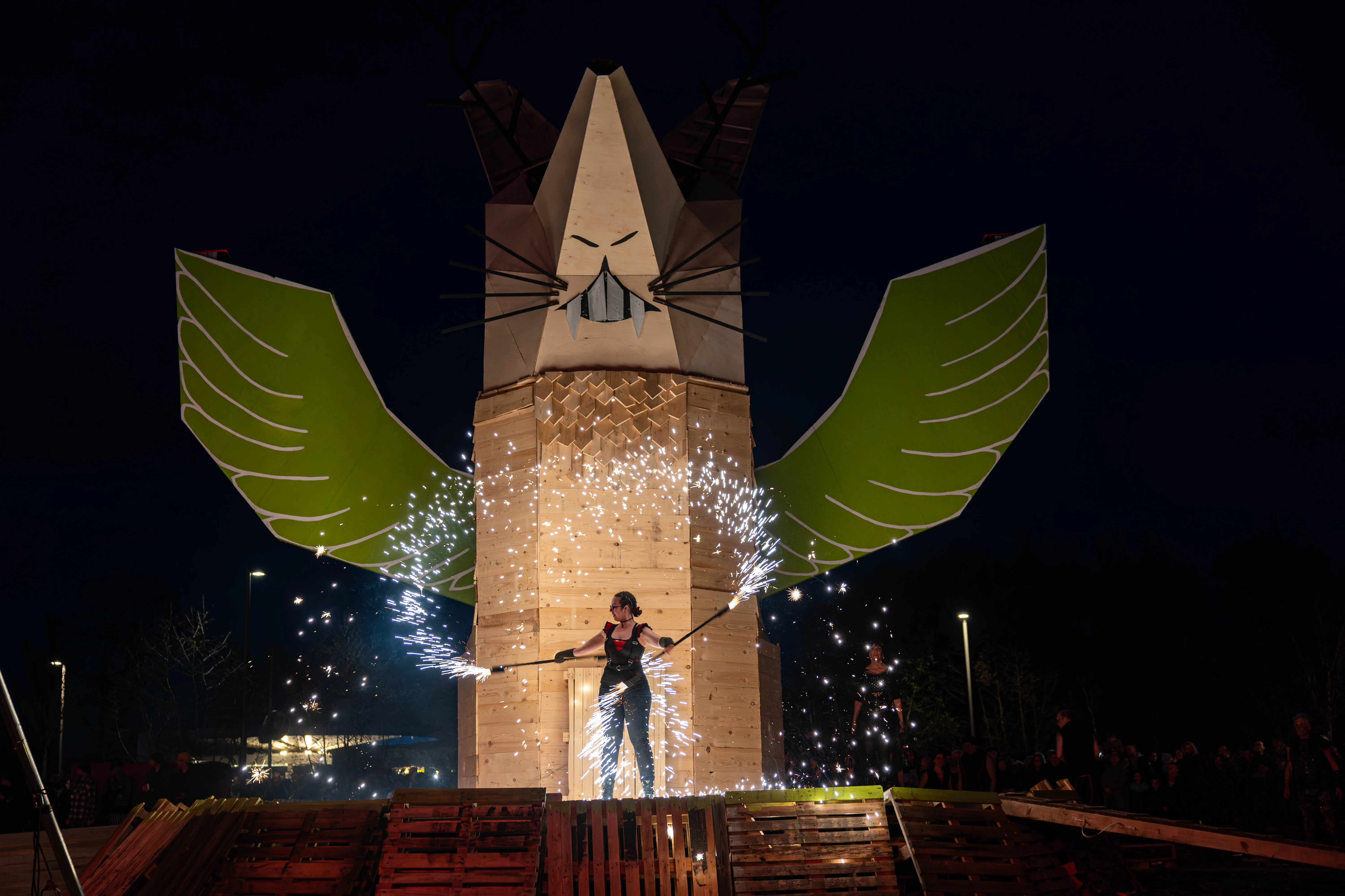 Person mit funkelnden Stäben vor einer großen, fuchsähnlichen Holzkonstruktion mit Flügeln bei einem Osterfeuer in der Nacht.