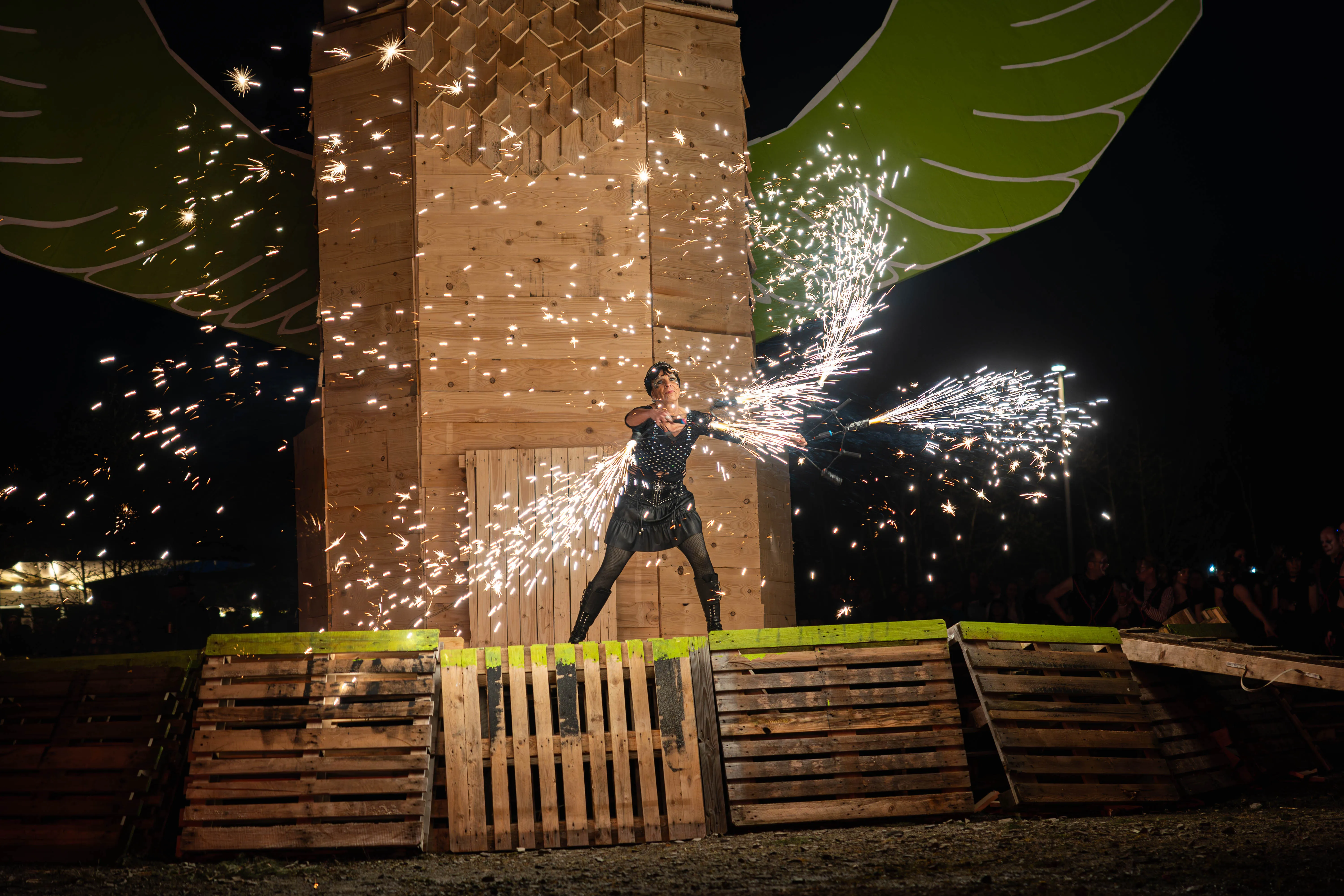Eine Person in dunkler Kleidung steht auf einer Holzkonstruktion vor einer großen Holzfassade und lässt Funken aus den Händen fliegen. Im Hintergrund sind grüne Flügelkonstruktionen und Zuschauer zu sehen.