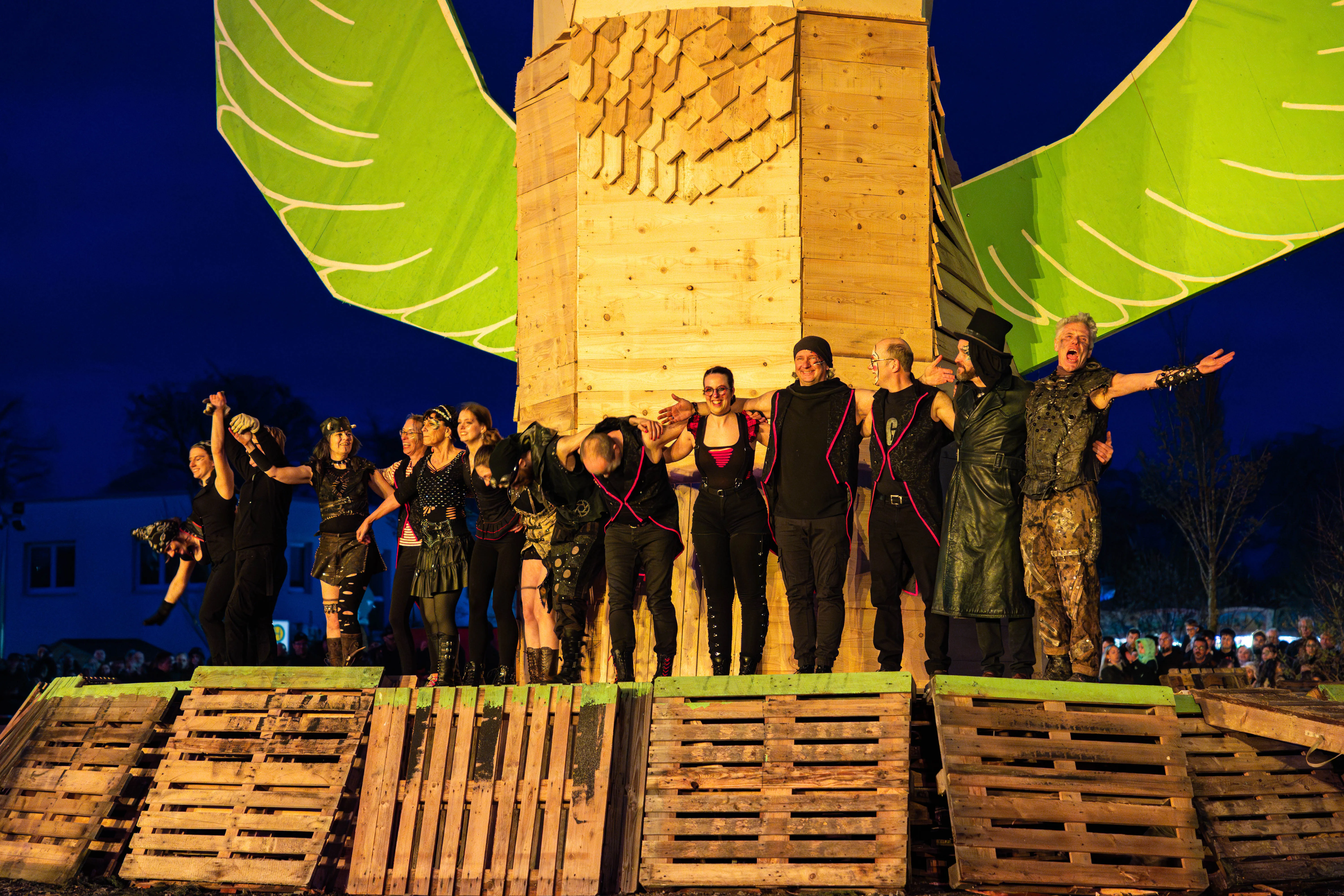 Eine Gruppe von Menschen in dunkler Kleidung steht auf Paletten vor einem großen, hölzernen Bauwerk mit leuchtend grünen Flügeln im Hintergrund. Im Vordergrund gestikuliert ein Mann mit erhobener Hand.