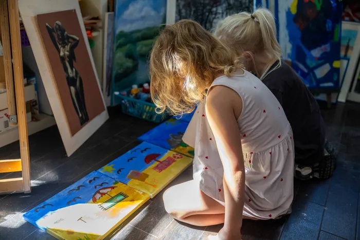 Zwei Kinder sitzen auf dem Boden und malen auf bunten Leinwänden in einem Kunstraum.