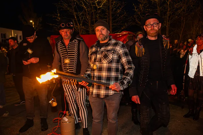 Mehrere Personen in fantasievoller Kleidung stehen um eine brennende Metallstange. Im Hintergrund sind weitere Menschen und ein Gebäude zu sehen.