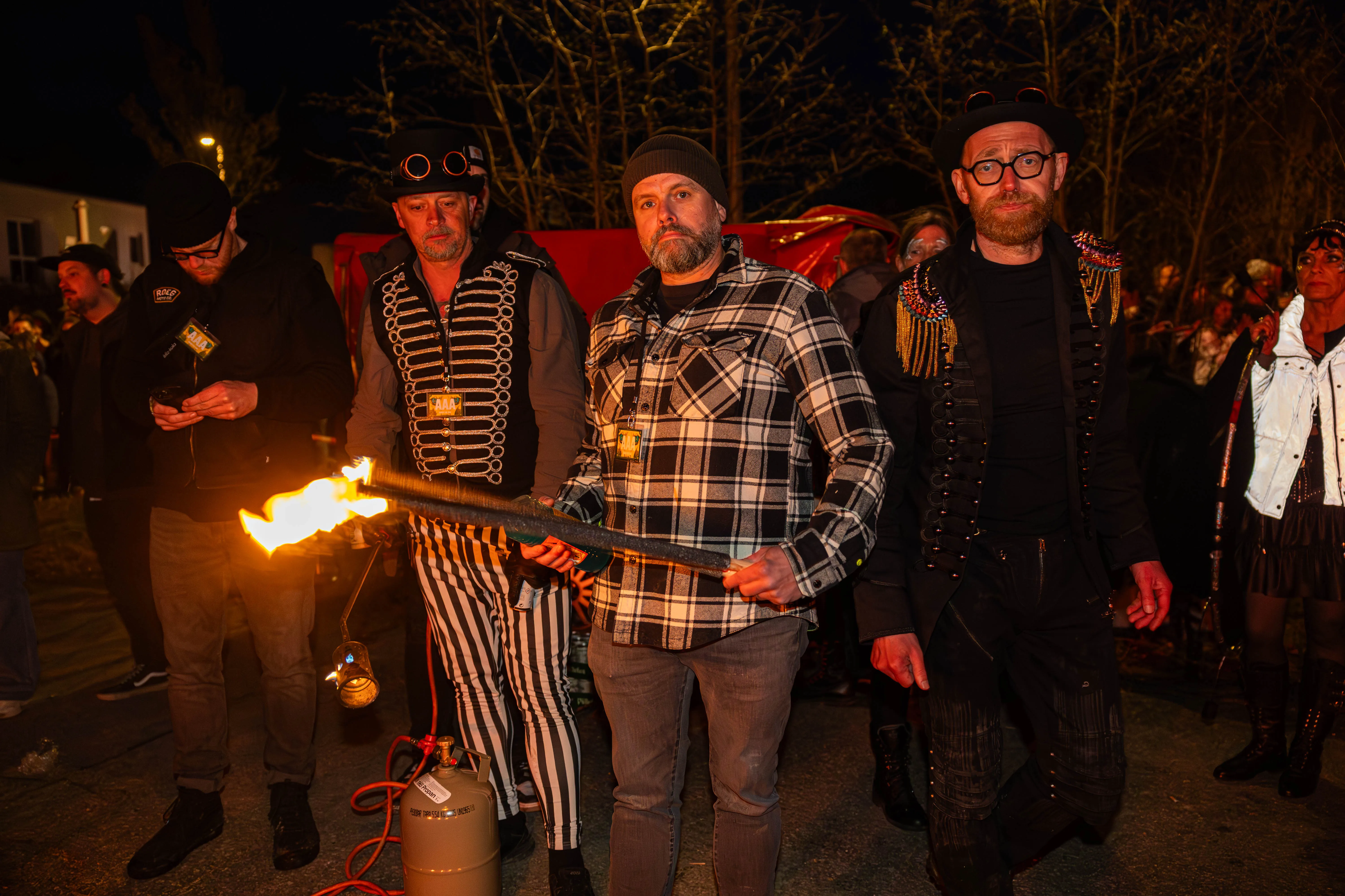 Mehrere Personen in fantasievoller Kleidung stehen um eine brennende Metallstange. Im Hintergrund sind weitere Menschen und ein Gebäude zu sehen.