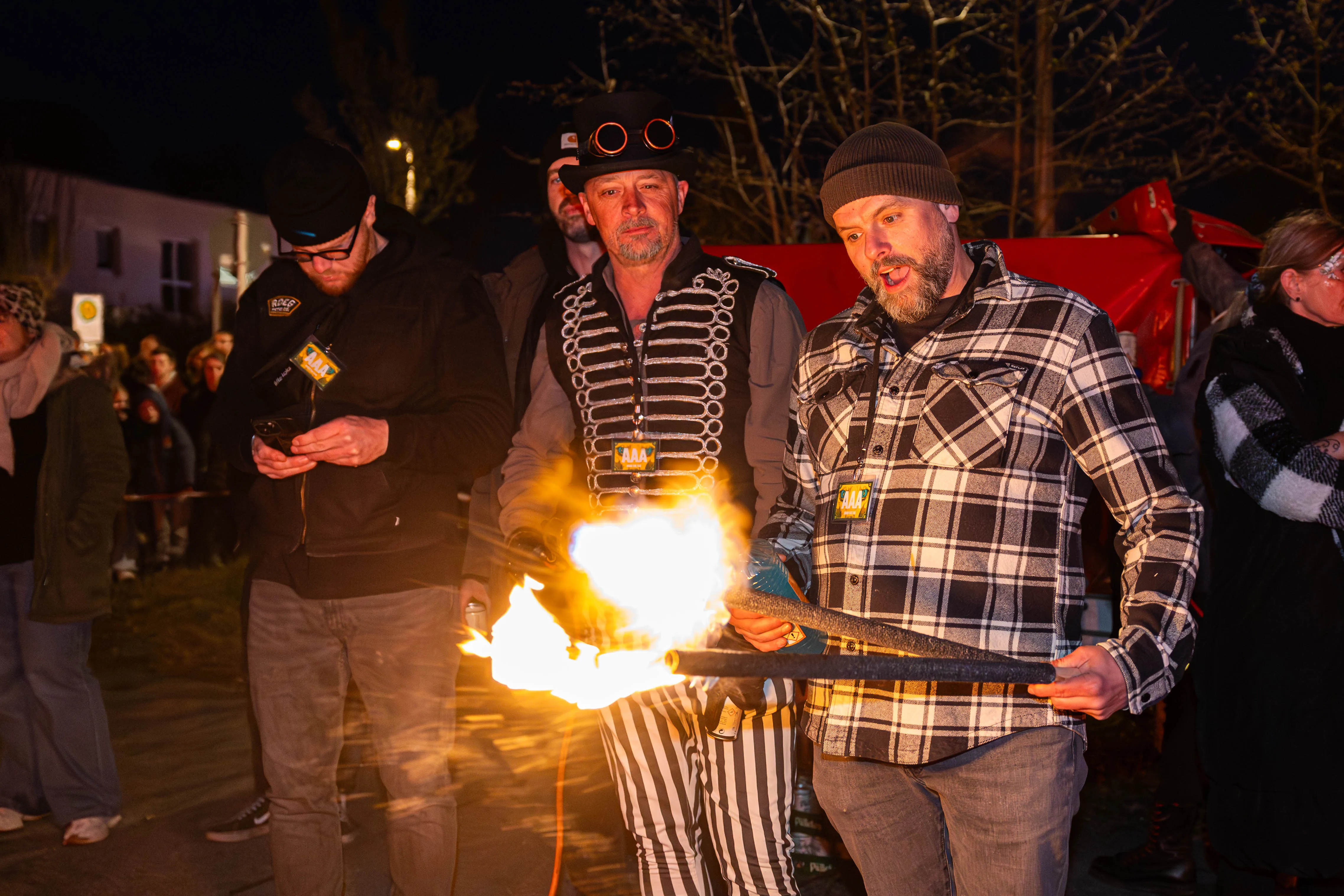 Drei Männer halten brennende Fackeln während eines Osterfeuers in Lüdenscheid. Im Hintergrund sind weitere Menschen und ein rotes Fahrzeug zu sehen.