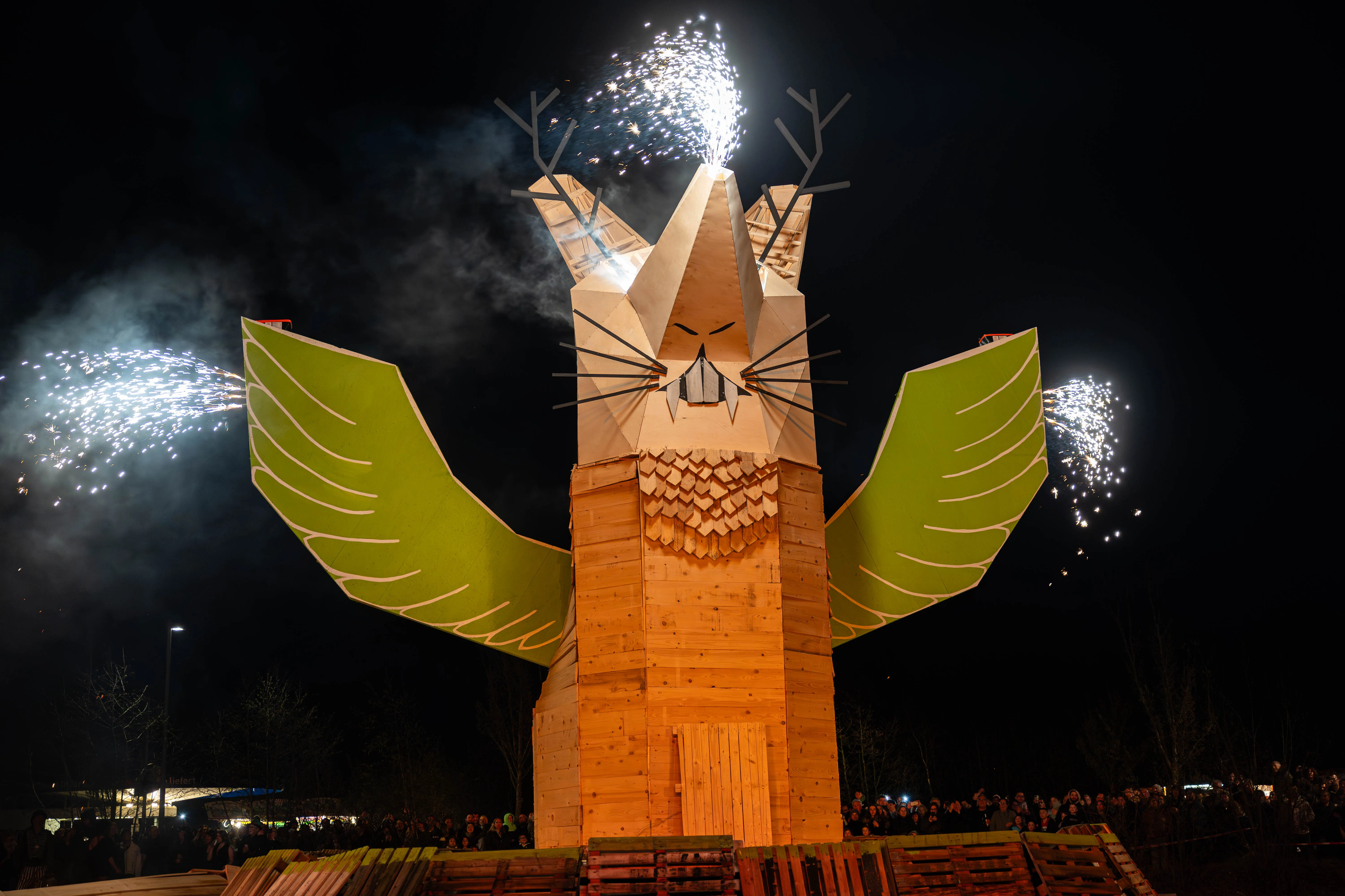 Große Holzskulptur eines Fuchses mit ausgebreiteten grünen Flügeln, während Feuerwerk in den Nachthimmel steigt. Im Hintergrund eine Menschenmenge.