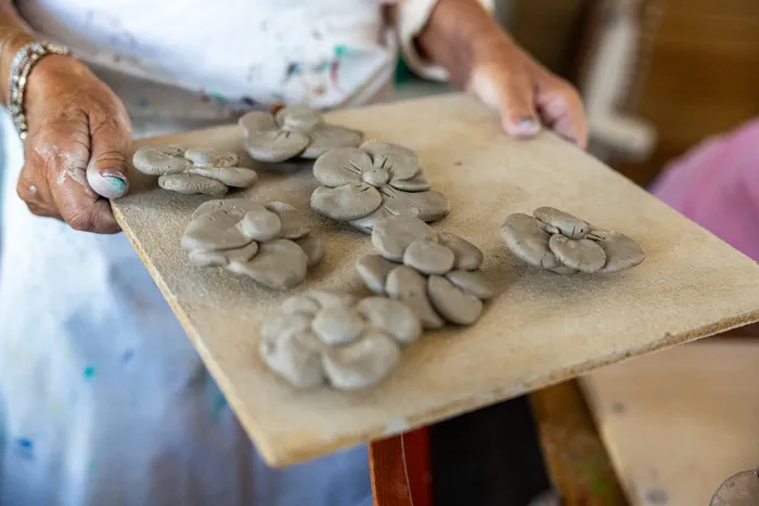 Hände halten ein Holzbrett mit mehreren kleinen, aus grauem Ton geformten Blumen.