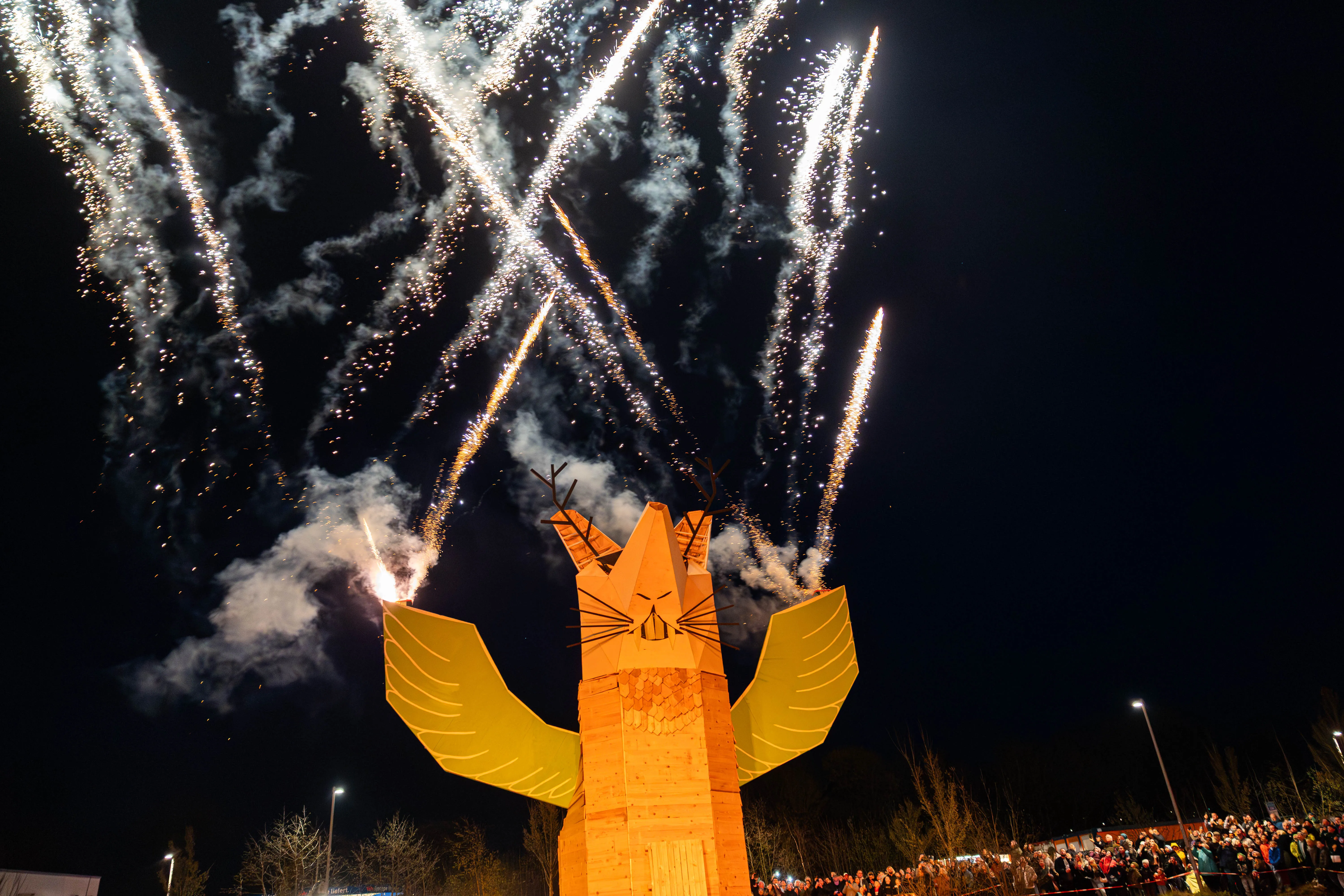 Großes Holz-Fuchs-Bauwerk mit ausgebreiteten Flügeln vor Feuerwerk bei Nacht. Im Hintergrund eine Menschenmenge.