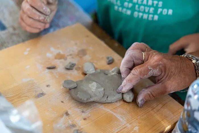 Mit Ton bedeckte Hände formen eine kleine Figur aus grauem Ton auf einem Holzbrett.