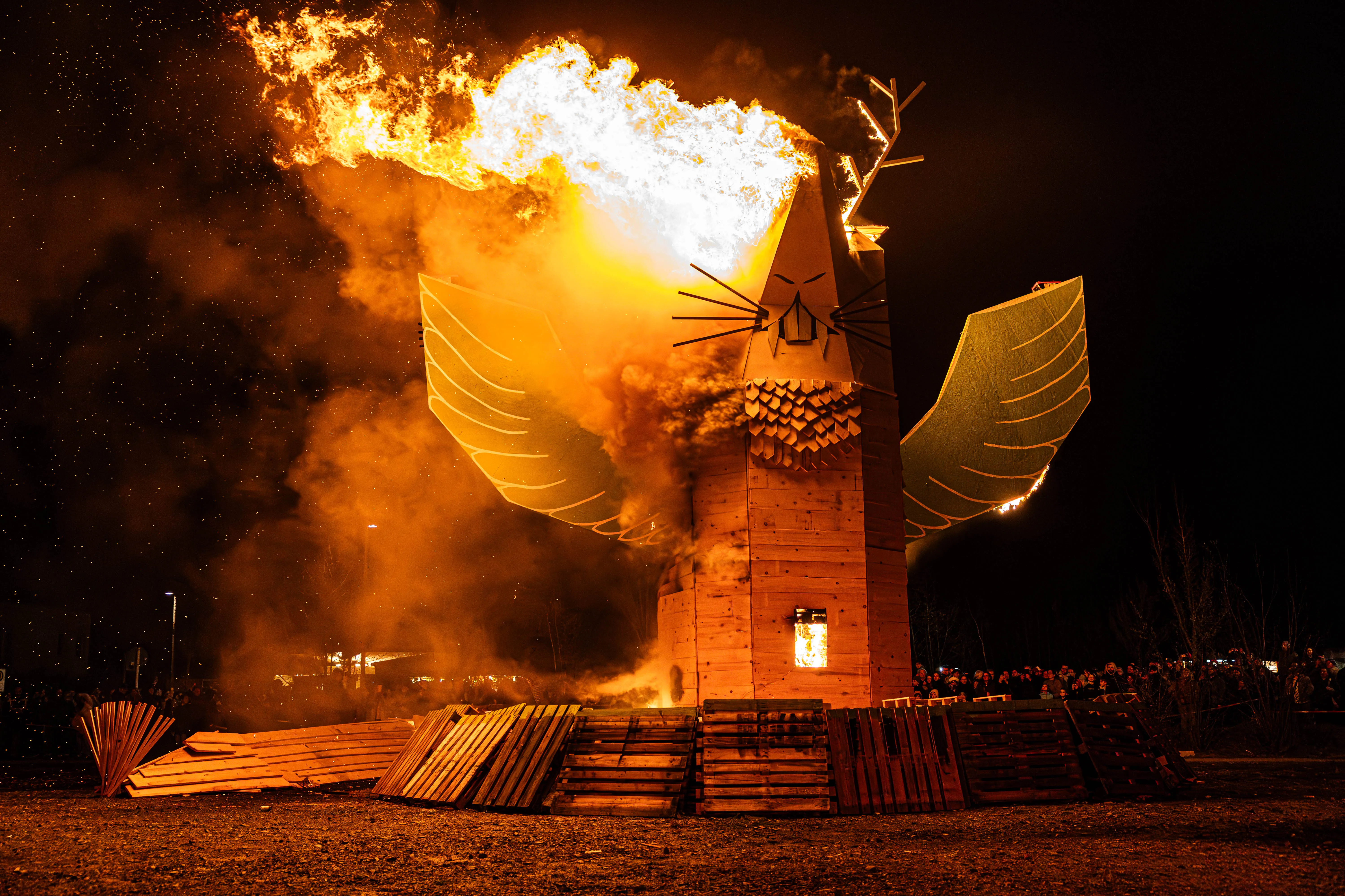 Großer brennender Holzfuchs in Flammen, umgeben von Rauch und Funken, vor dunklem Nachthimmel. Im Hintergrund verschwommene Menschenmenge.