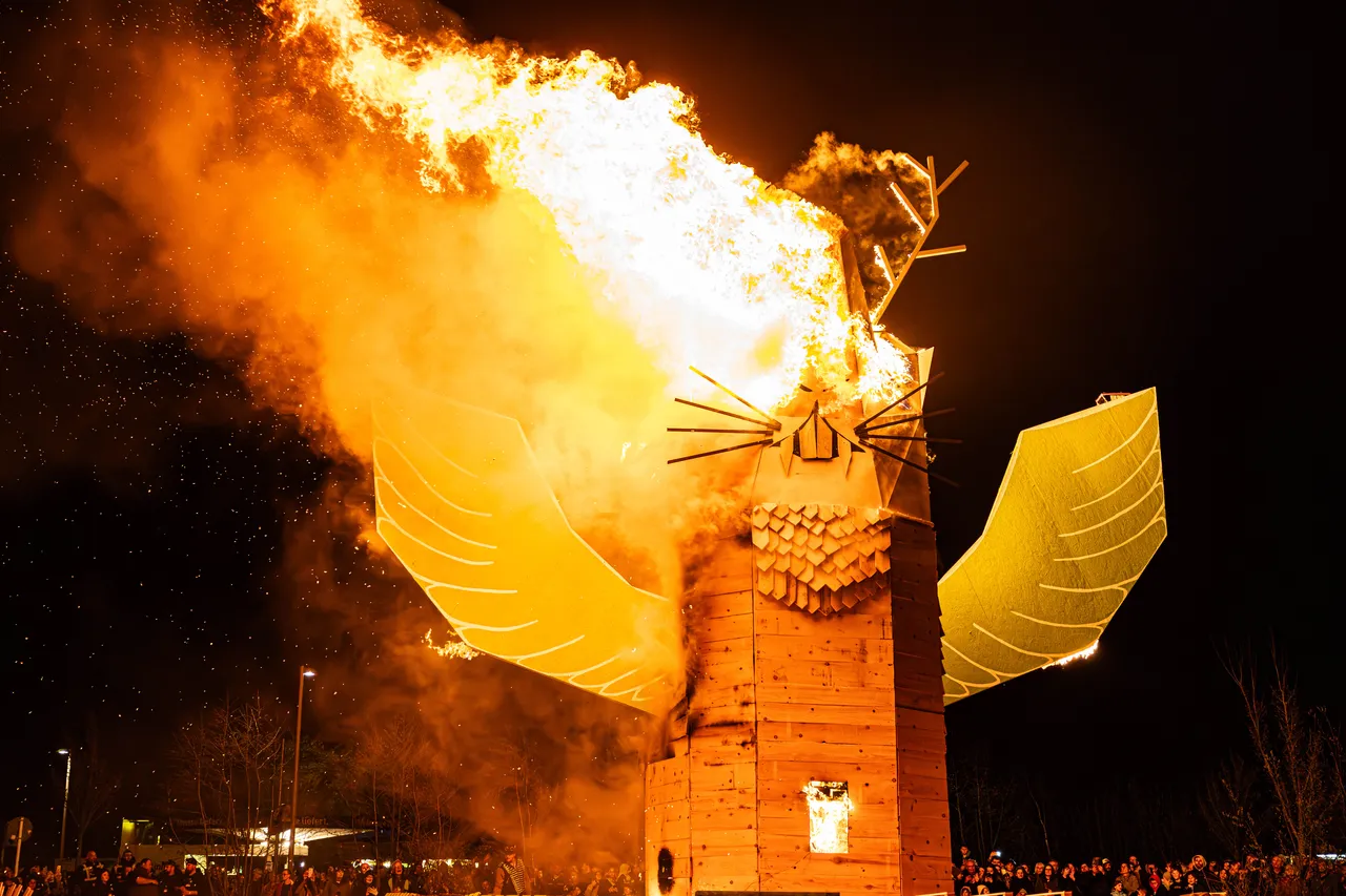 Großer, brennender Holzsteg in Form eines Fuchses mit ausgebreiteten Flügeln vor dunklem Nachthimmel. Im Hintergrund verschwommene Menschenmenge.