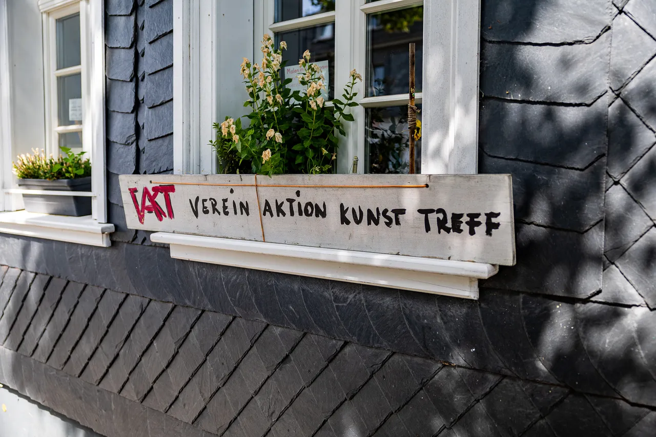 Ein hölzerner Blumenkasten mit der Aufschrift 'VAKT VEREIN AKTION KUNST TREFF' vor einem Fenster an einer dunklen Schieferfassade.