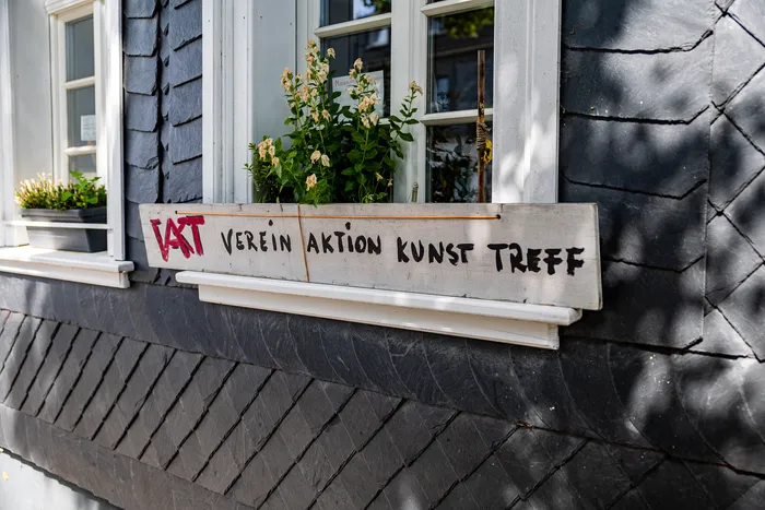 Ein hölzerner Blumenkasten mit der Aufschrift 'VAKT VEREIN AKTION KUNST TREFF' vor einem Fenster an einer dunklen Schieferfassade.