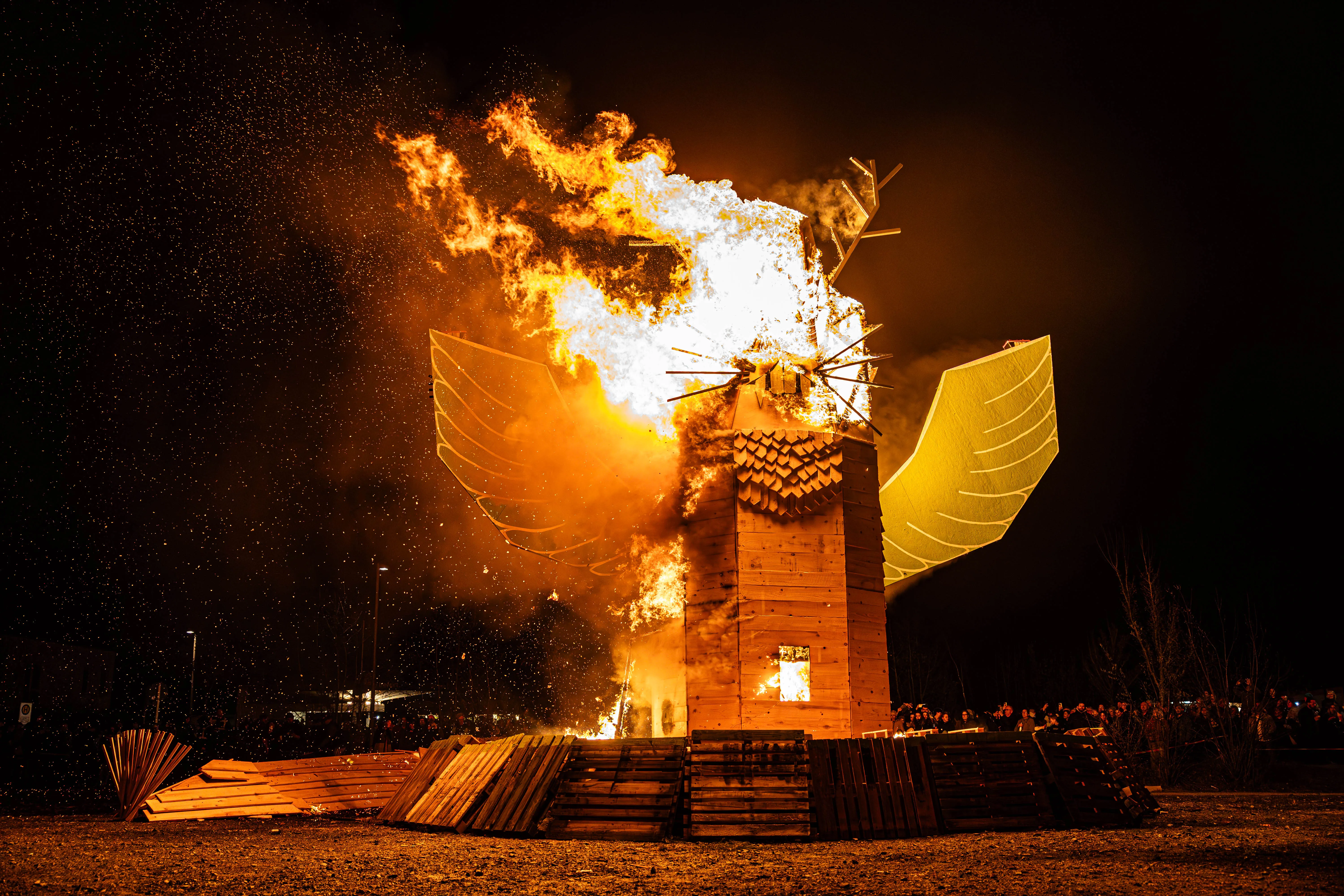 Großer, brennender Holzadler steht auf einem Podest im Freien, umgeben von Zuschauern im Dunkeln. Funken fliegen auf und beleuchten die Szene.