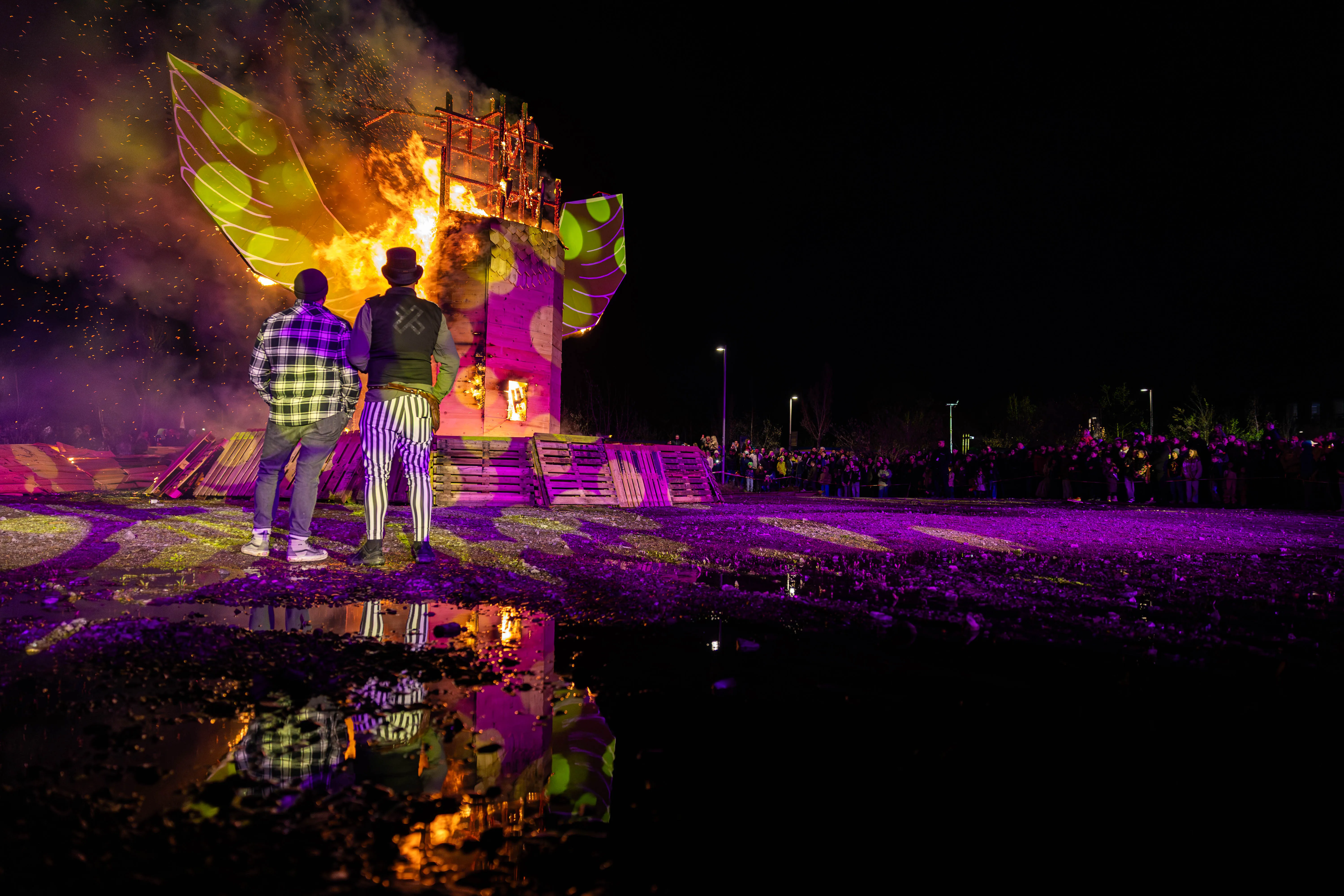 Zwei Personen stehen dem brennenden Holzkonstrukt mit Flügeln auf einem Feld zu, im Hintergrund eine Menschenmenge und violettes Licht.