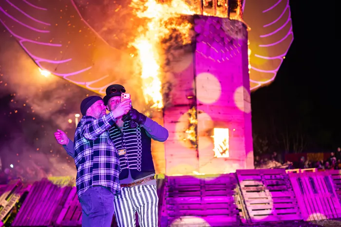 Zwei Personen in Kostümen umarmen sich vor einem brennenden Holzgebäude bei einem Osterfeuer und machen ein Selfie mit einem Smartphone.