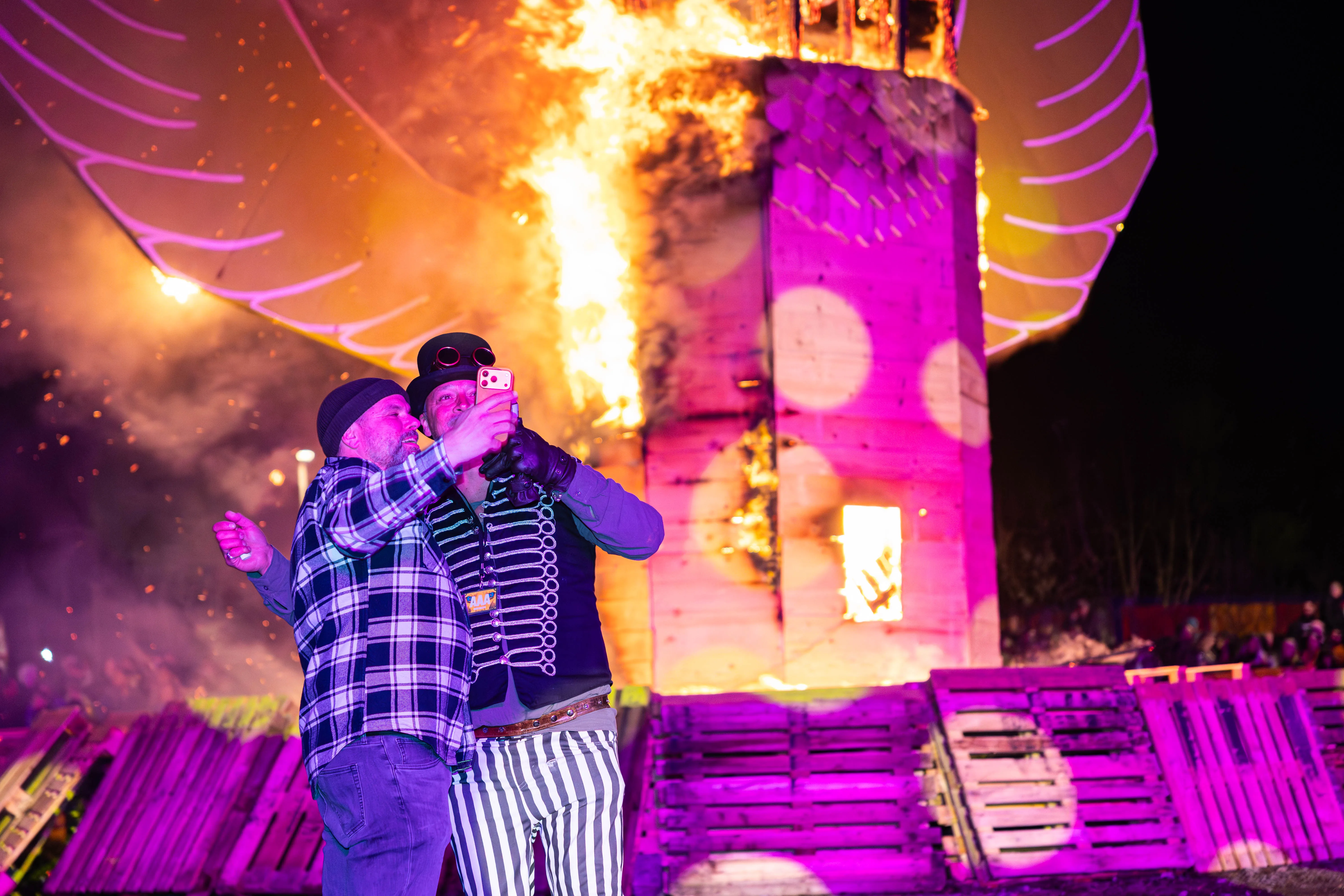 Zwei Personen in Kostümen umarmen sich vor einem brennenden Holzgebäude bei einem Osterfeuer und machen ein Selfie mit einem Smartphone.
