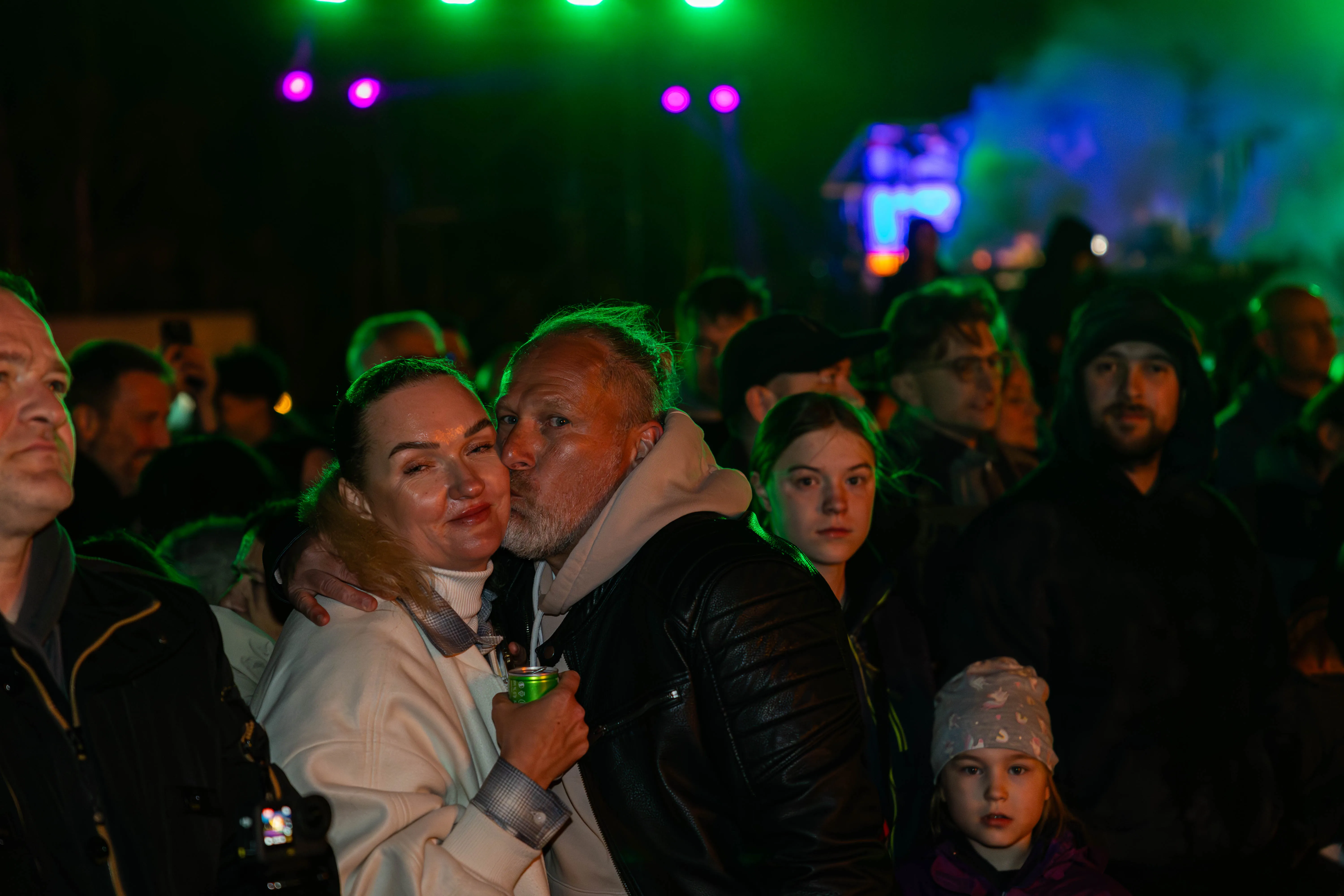 Ein Paar umarmt sich eng vor einer Menschenmenge bei einem Osterfeuer mit grünem und violettem Licht im Hintergrund.