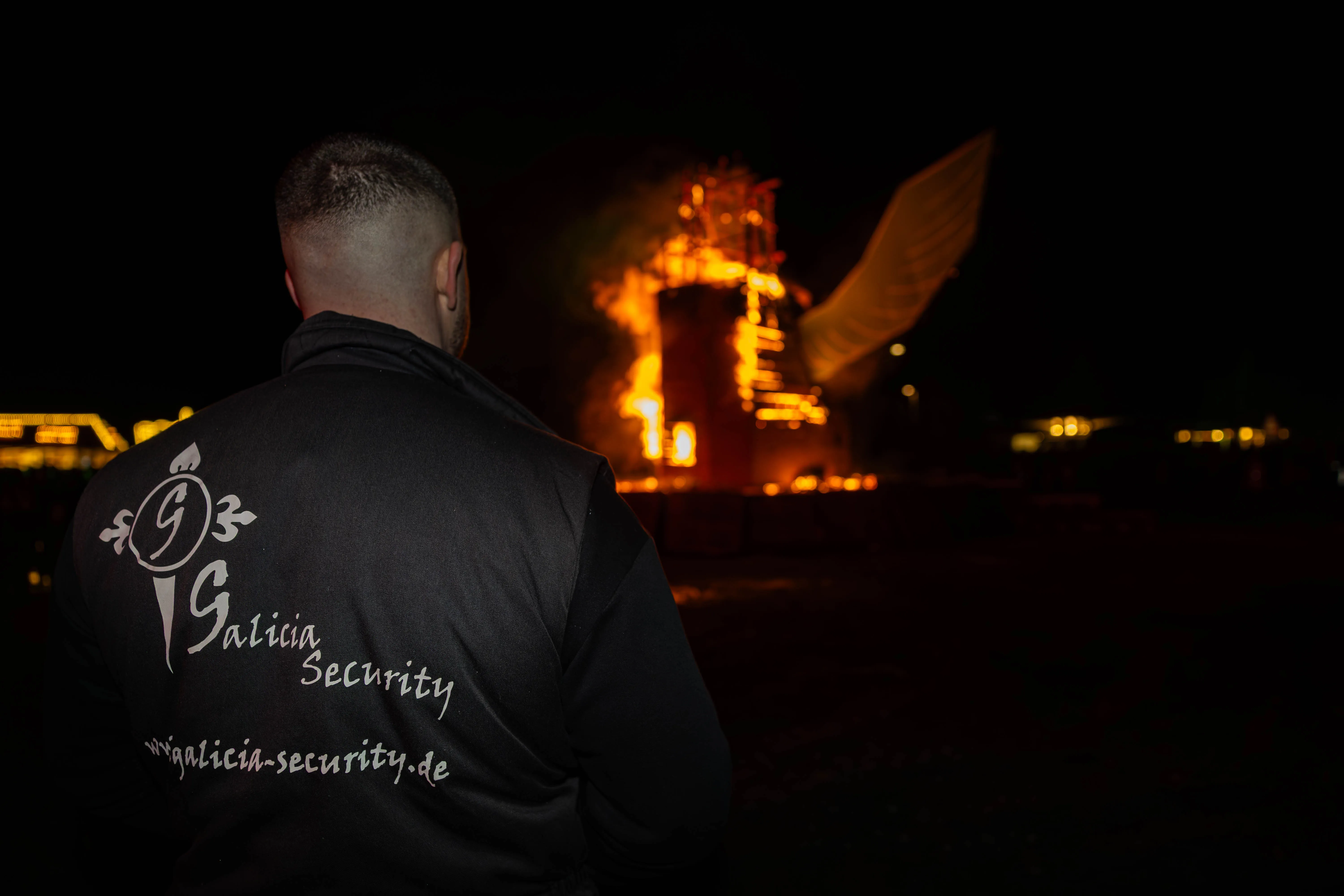 Sicherheitsdienstmitarbeiter mit dem Rücken zur Kamera, im Hintergrund ein großes Osterfeuer mit einer brennenden Figur in Form eines Fuchses.
