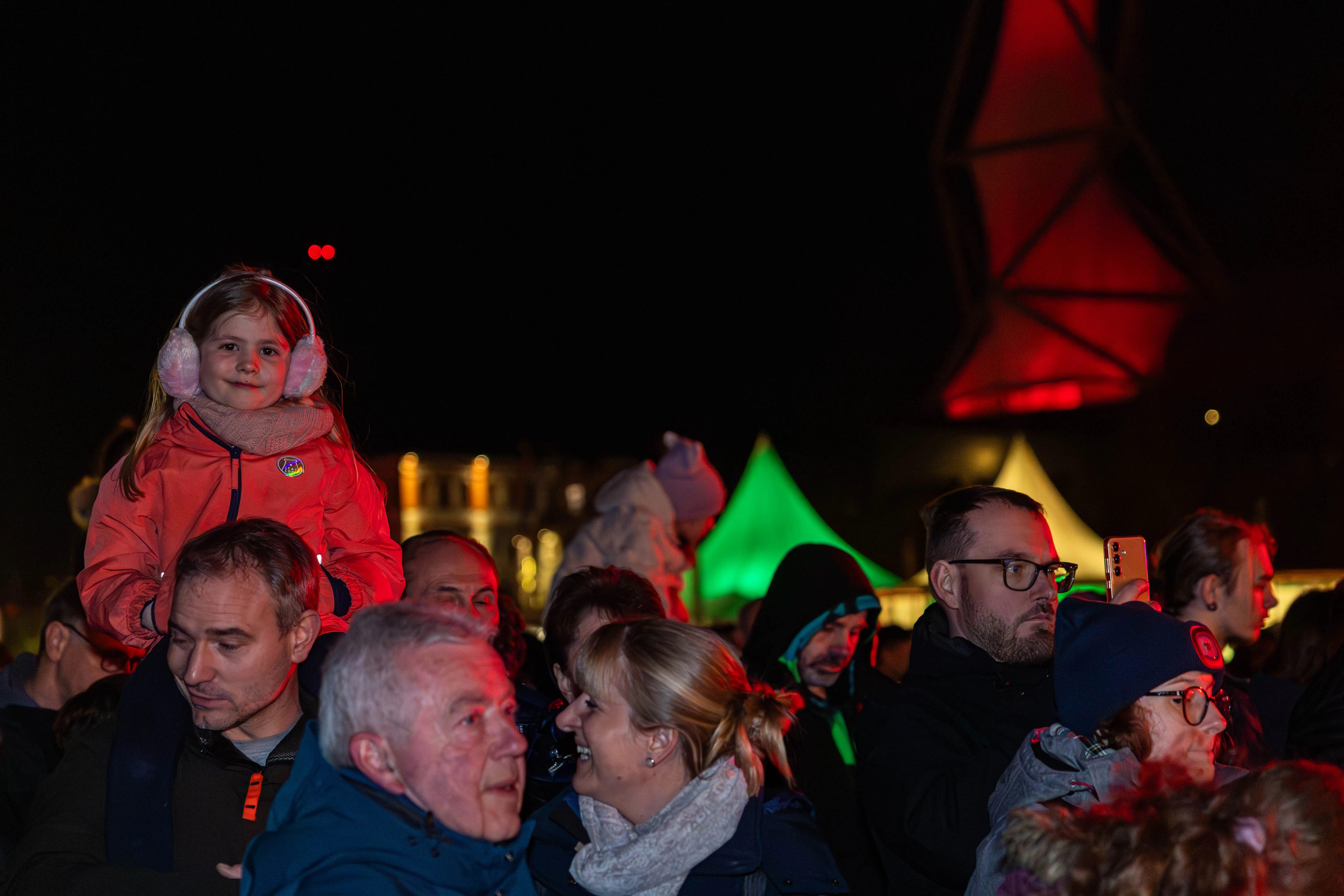 Ein kleines Mädchen mit Kopfhörern sitzt auf den Schultern eines Mannes vor einer Menschenmenge, die ein Feuerwerk betrachtet. Im Hintergrund leuchten rote und grüne Lichter.