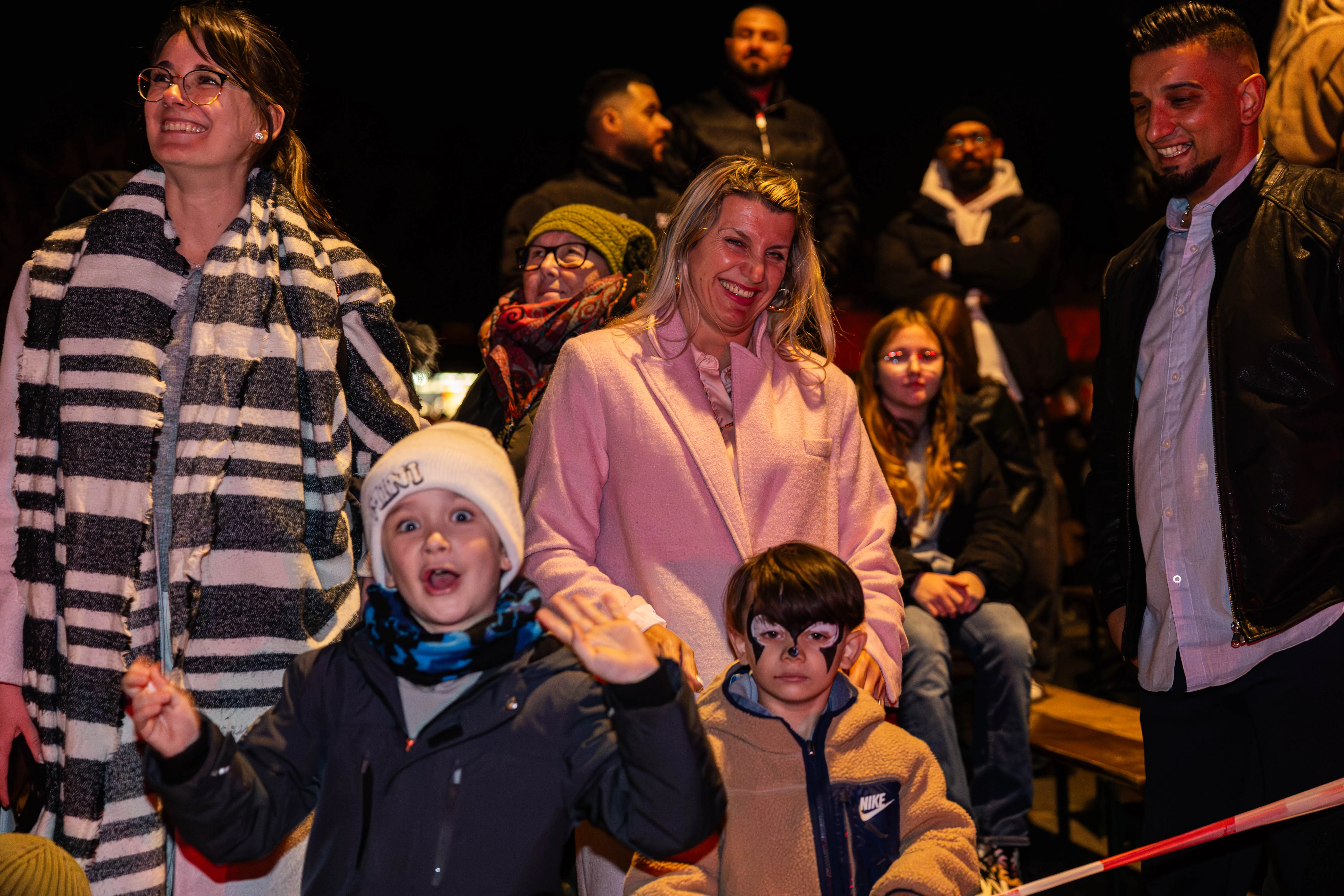 Zuschauer, darunter Kinder, lächeln und jubeln bei einer Veranstaltung im Freien. Im Hintergrund sind weitere Menschen und ein rotes Absperrband zu sehen.