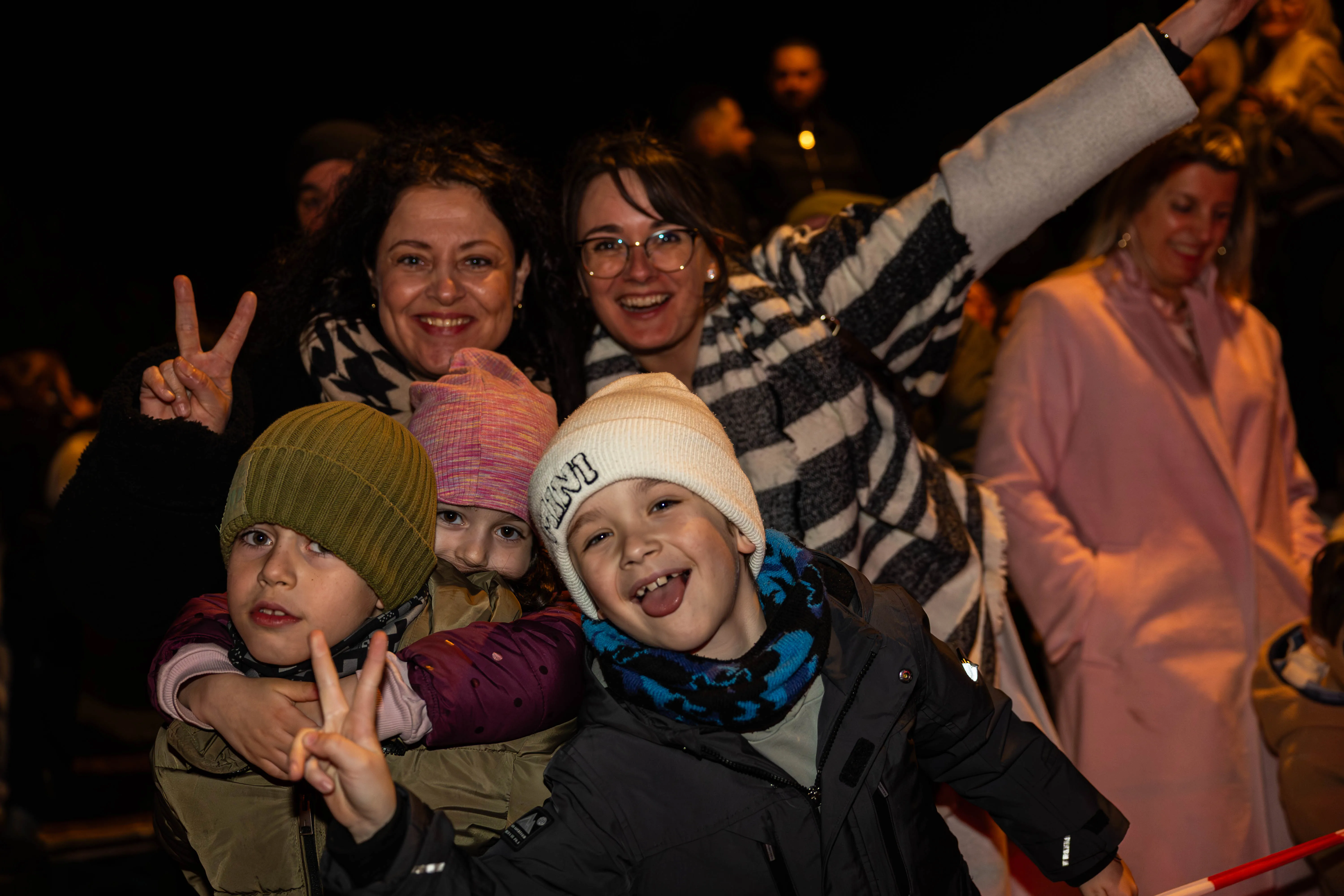 Mehrere Personen, darunter Kinder mit Mützen und Frauen, lächeln in die Kamera und zeigen das Friedenszeichen. Im Hintergrund sind weitere Menschen und ein dunkler Nachthimmel zu sehen.