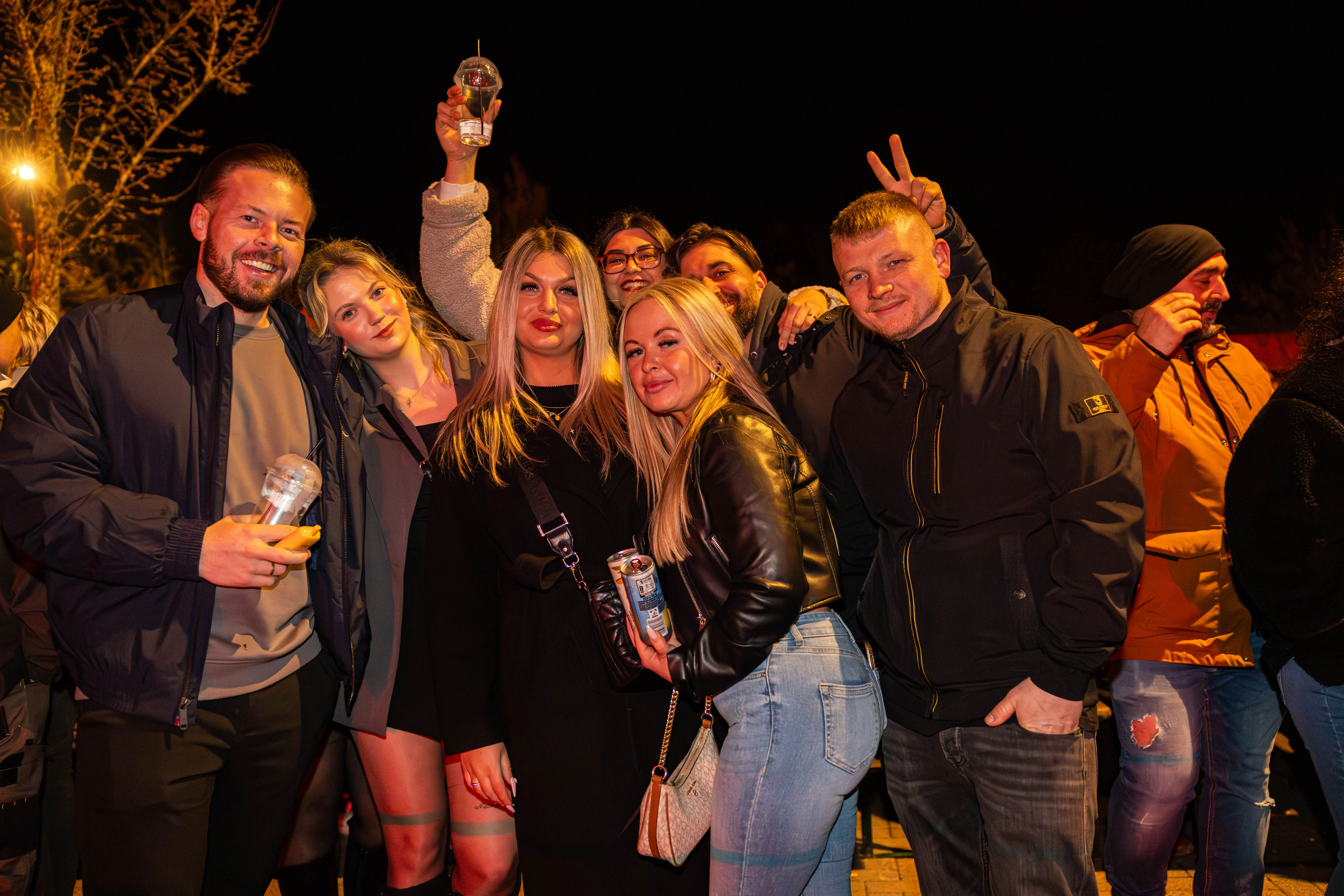 Eine Gruppe von jungen Erwachsenen posiert eng zusammen vor dunklem Hintergrund, einige halten Getränke in der Hand. Im Hintergrund sind weitere Menschen und Lichter zu sehen.