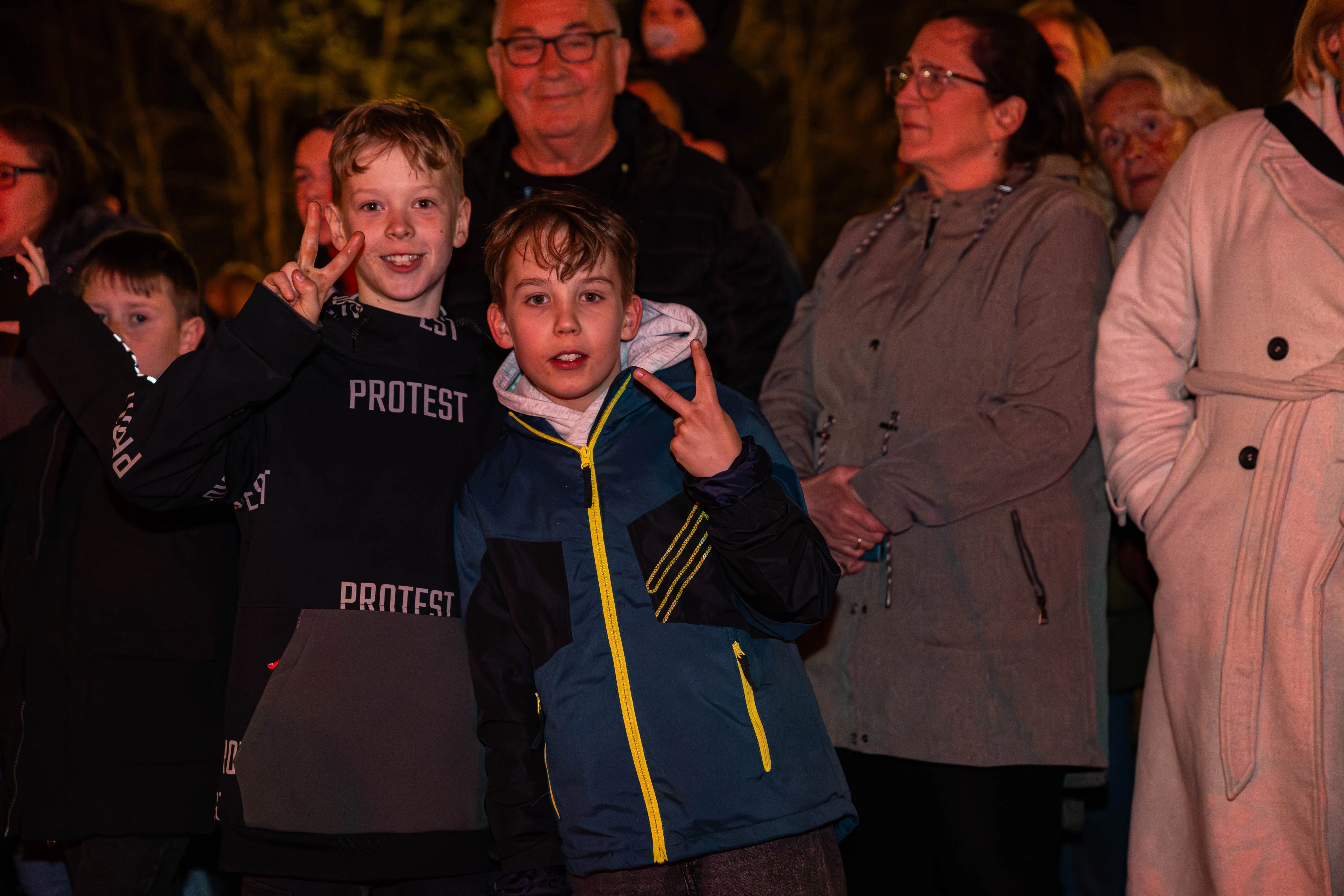 Zwei Jungen im Vordergrund machen das Victory-Zeichen, während im Hintergrund Menschen ein Osterfeuer beobachten.