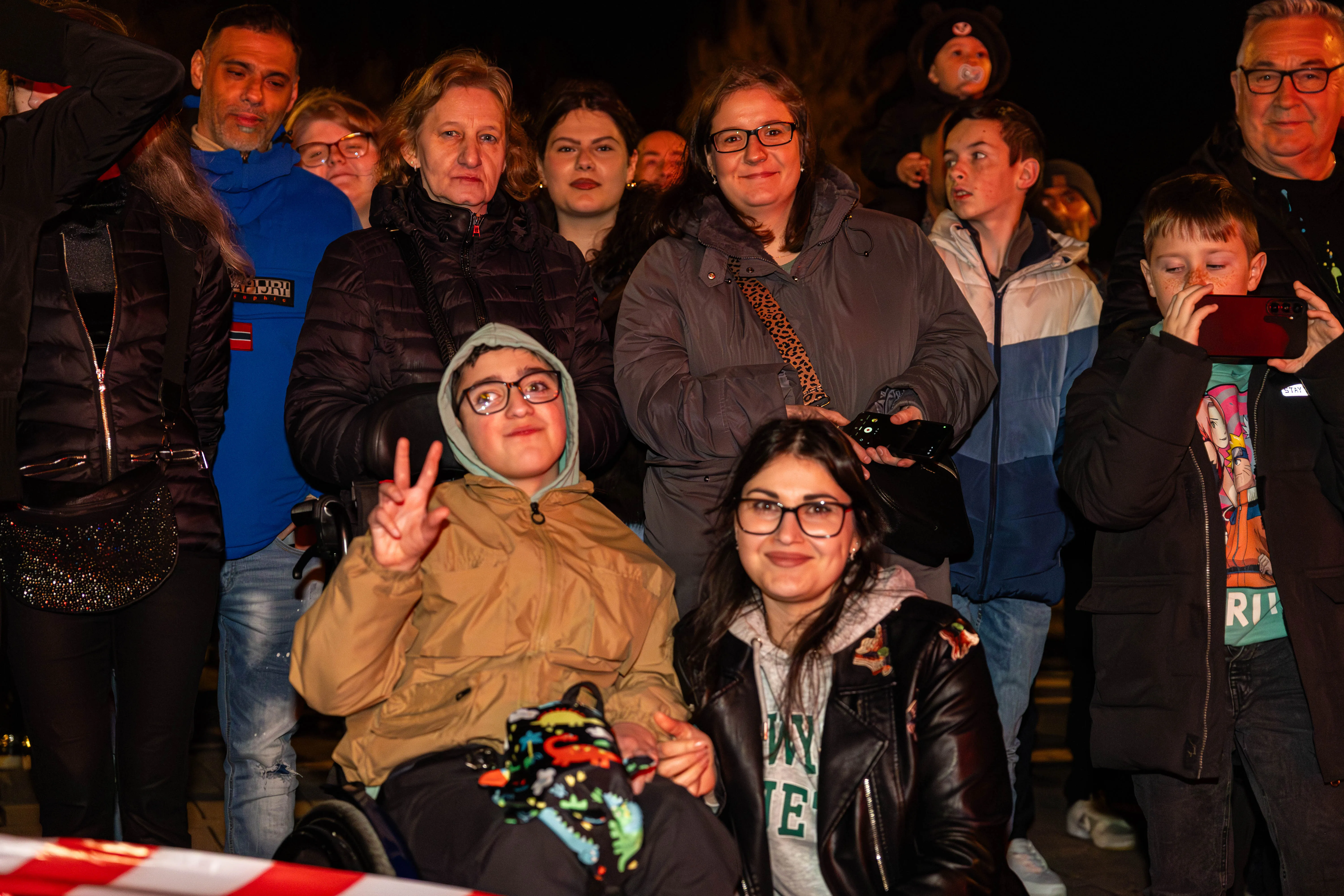 Menschengruppe vor dunklem Hintergrund, einige zeigen Friedenszeichen. Im Vordergrund eine Frau im Rollstuhl und eine junge Frau mit Brille.