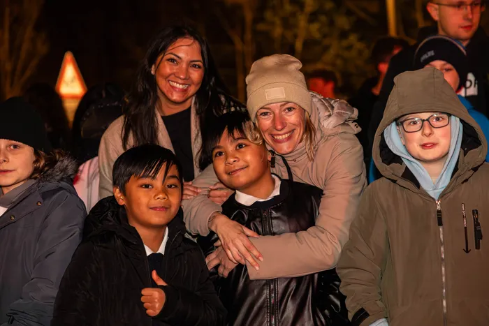 Mehrere Menschen lächeln in die Kamera, darunter zwei Frauen und drei Kinder unterschiedlicher Herkunft. Im Hintergrund sind weitere Personen und ein Feuer zu erkennen.