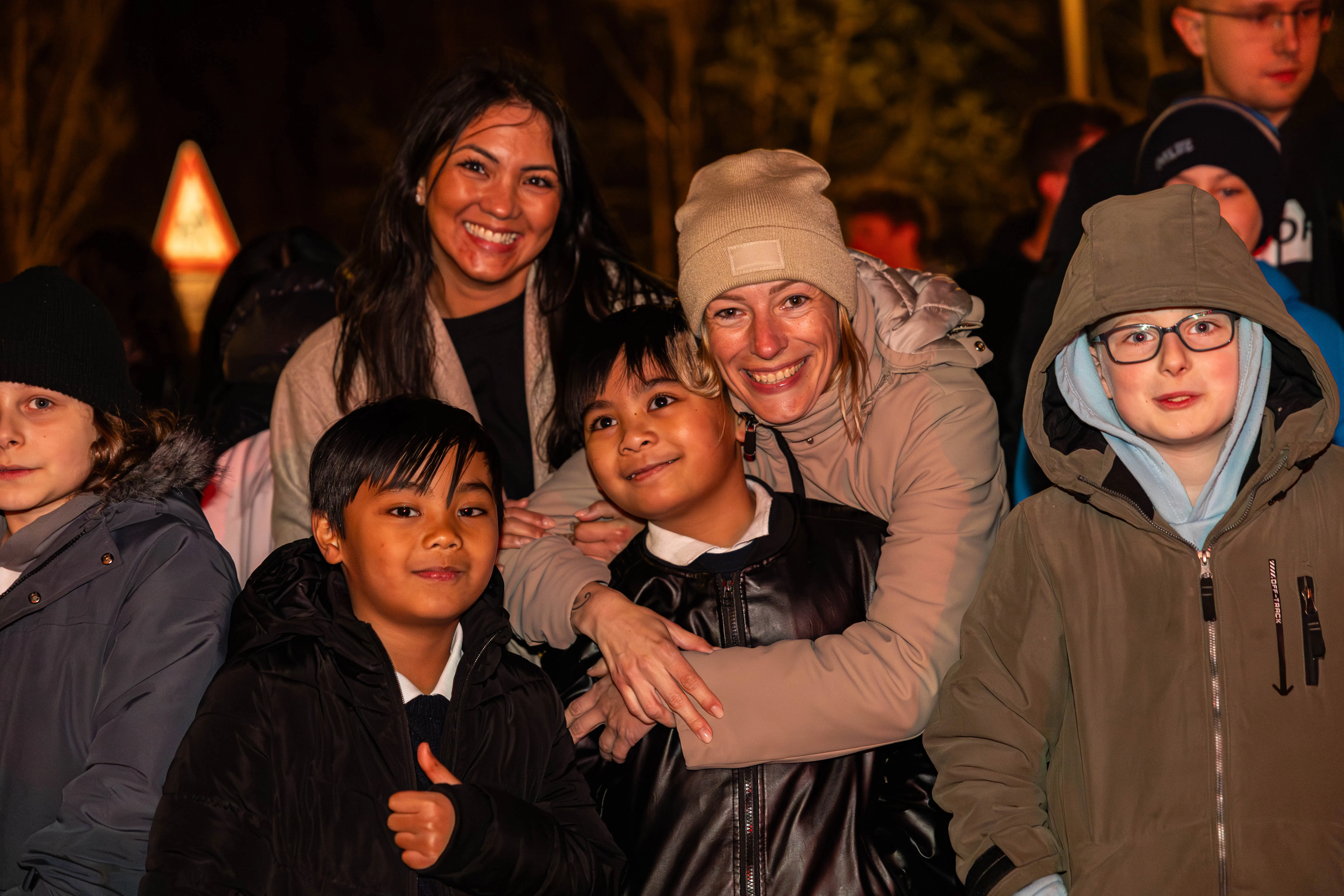 Mehrere Menschen lächeln in die Kamera, darunter zwei Frauen und drei Kinder unterschiedlicher Herkunft. Im Hintergrund sind weitere Personen und ein Feuer zu erkennen.