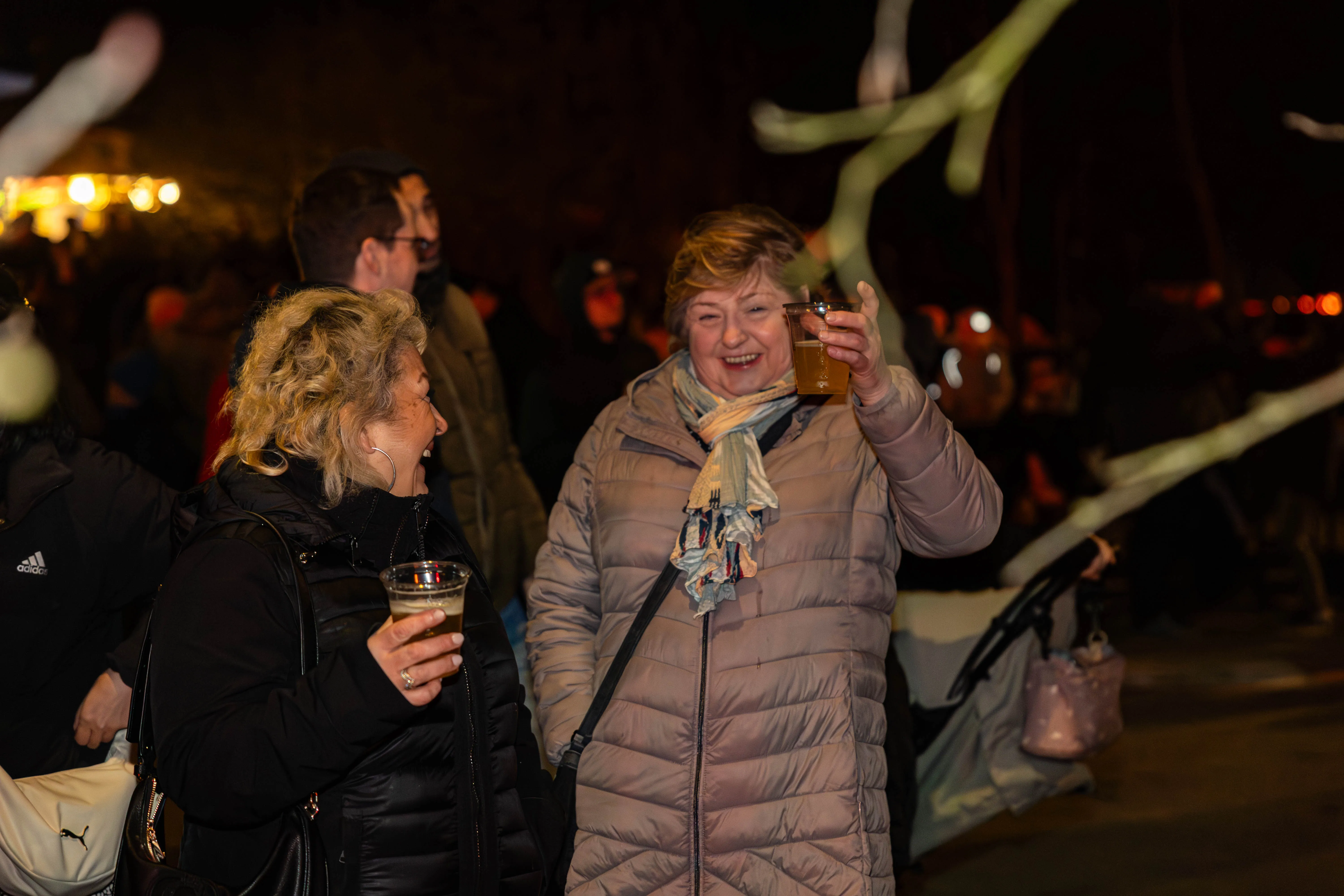 Zwei Frauen mit Getränken im Glas lächeln sich auf einem belebten Osterfeuer zu, im Hintergrund weitere Menschen und Lichtinstallationen.