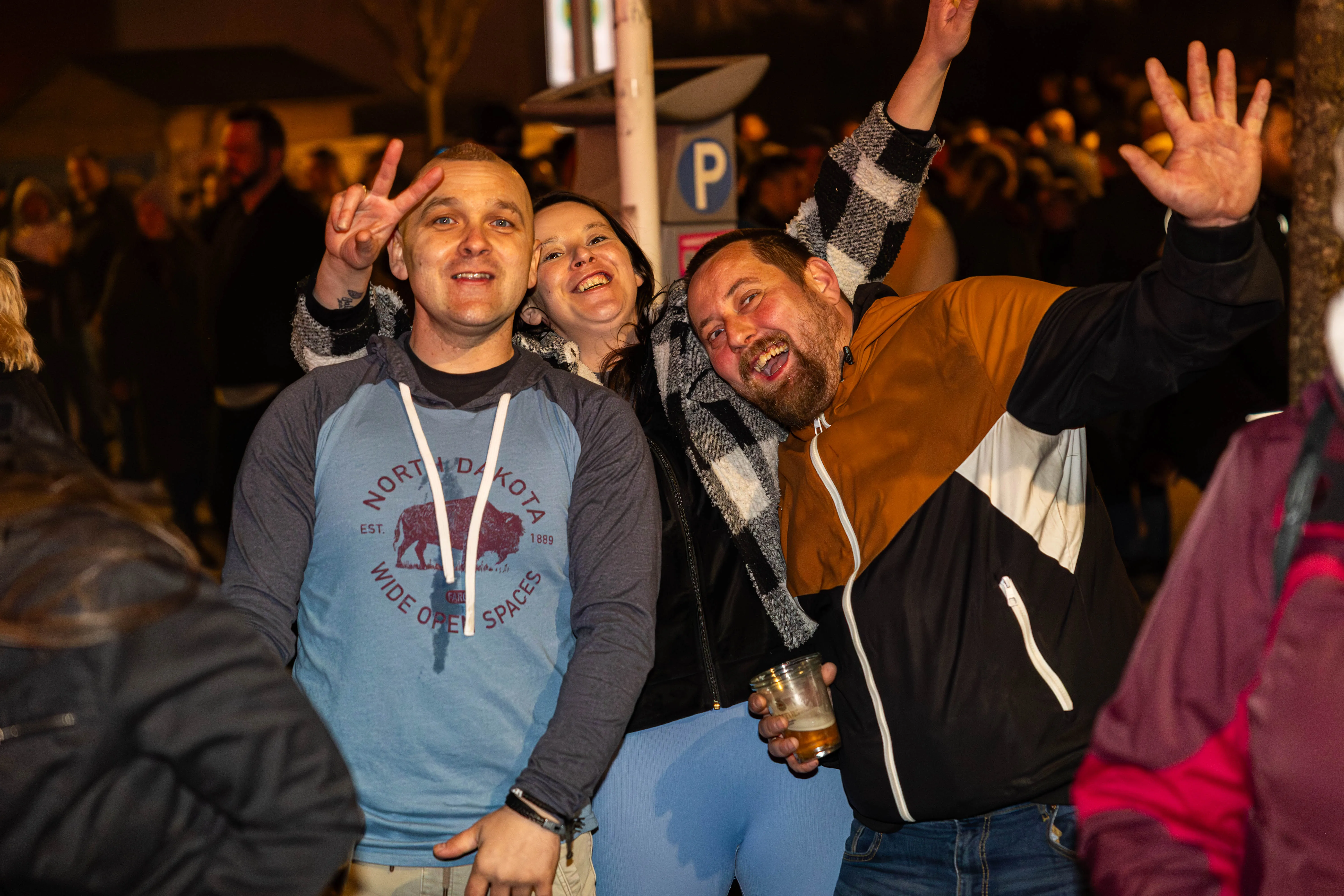Drei Menschen jubeln und lächeln in die Kamera, während sie inmitten einer Menschenmenge stehen. Einer der Männer hält ein Bierglas in der Hand.