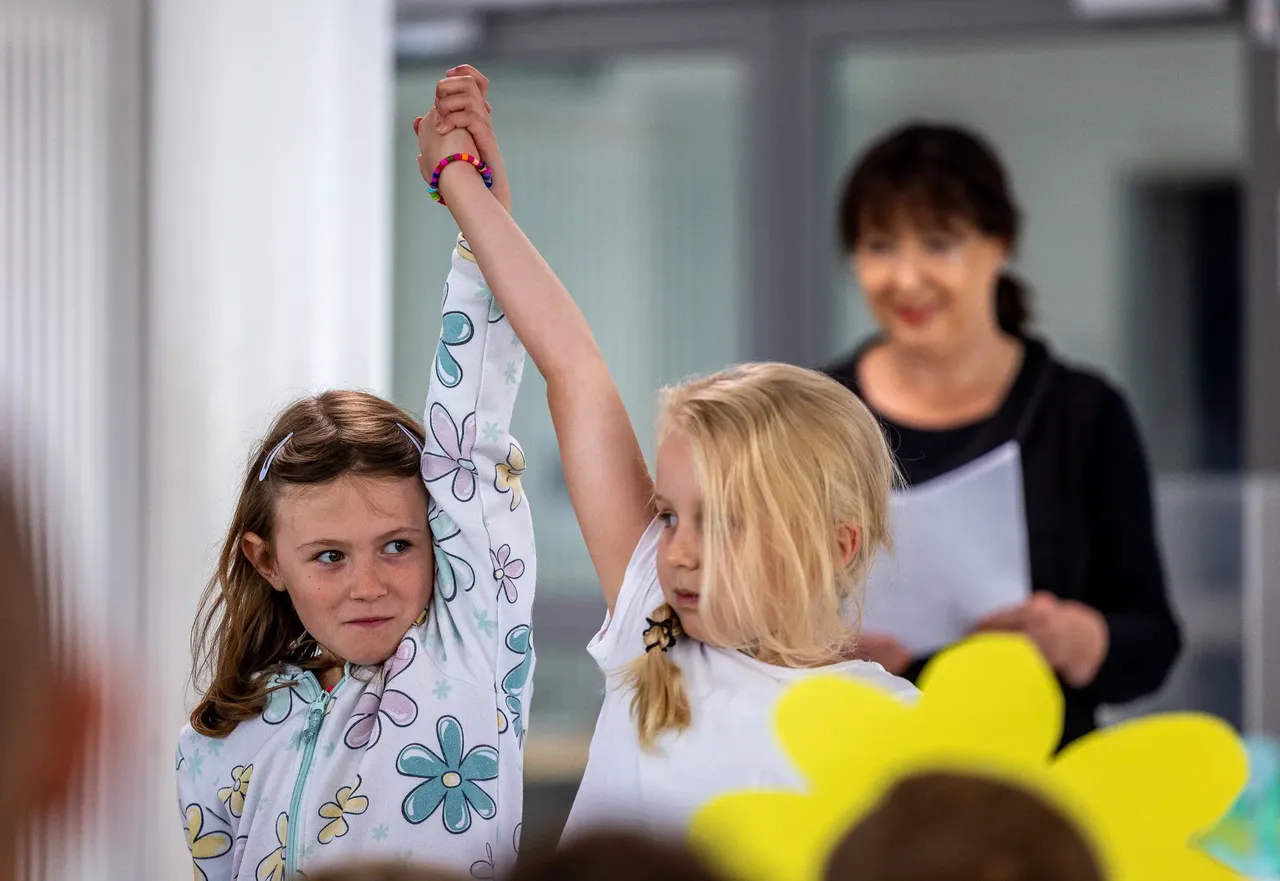 Zwei Mädchen bei einer Theateraufführung, wobei ein Mädchen die Hand des anderen Mädchens hochhält.