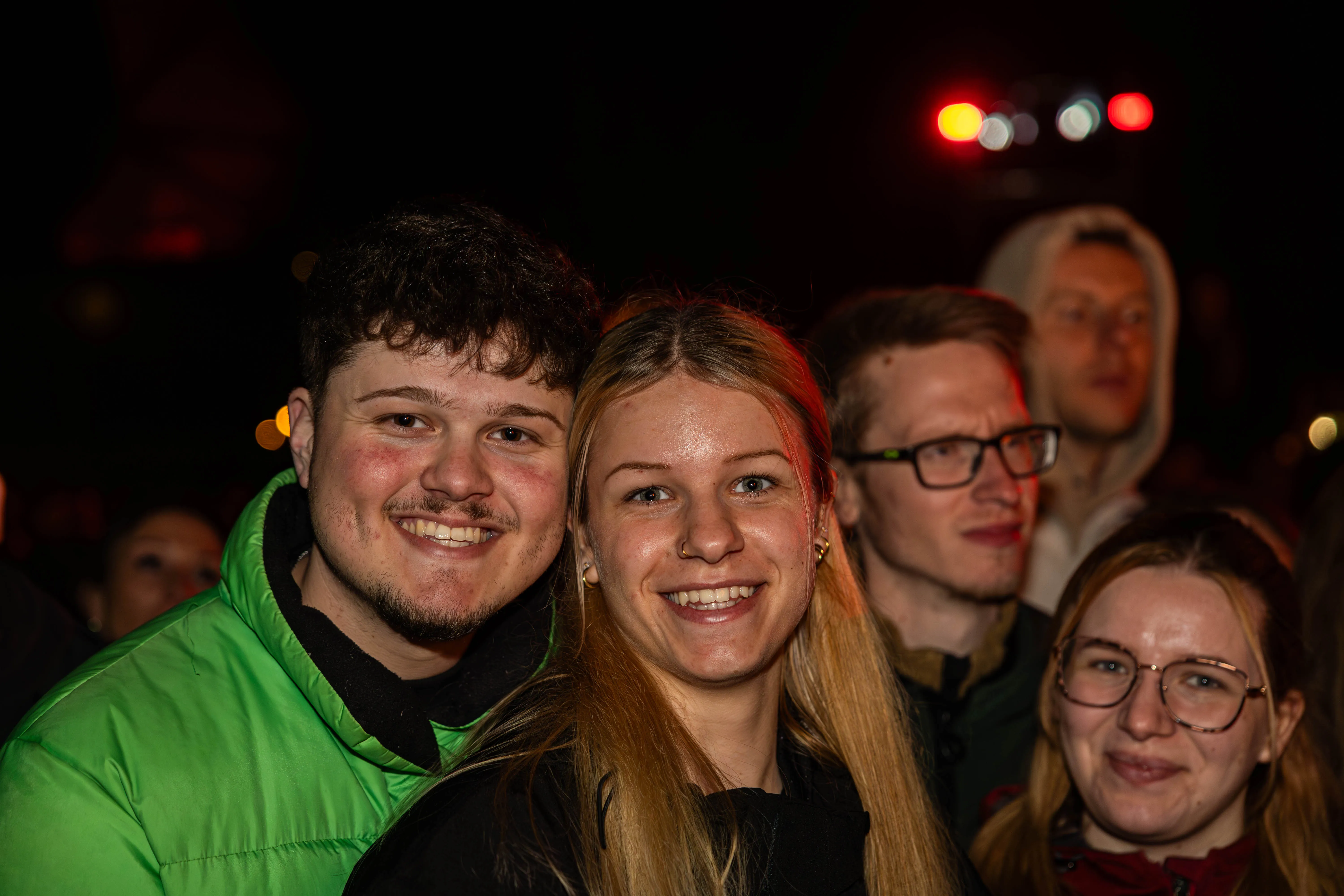 Nahaufnahme von lächelnden jungen Erwachsenen vor verschwommener Menschenmenge und rotem Licht bei einer Veranstaltung im Freien.