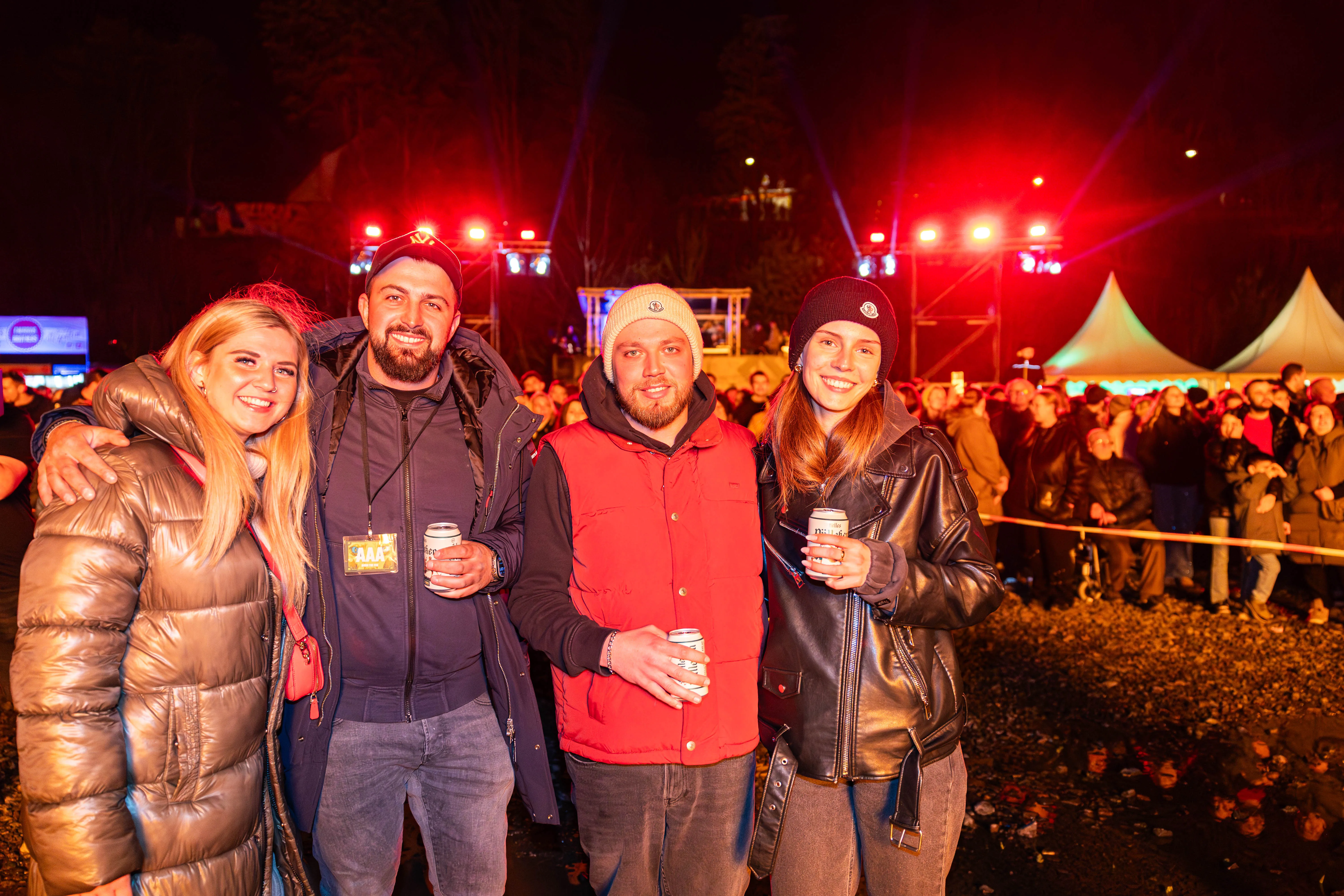 Vier junge Erwachsene lächeln in die Kamera, umgeben von einer Menschenmenge vor einer Bühne mit rotem Licht. Im Hintergrund sind Zelte und Bäume zu sehen.