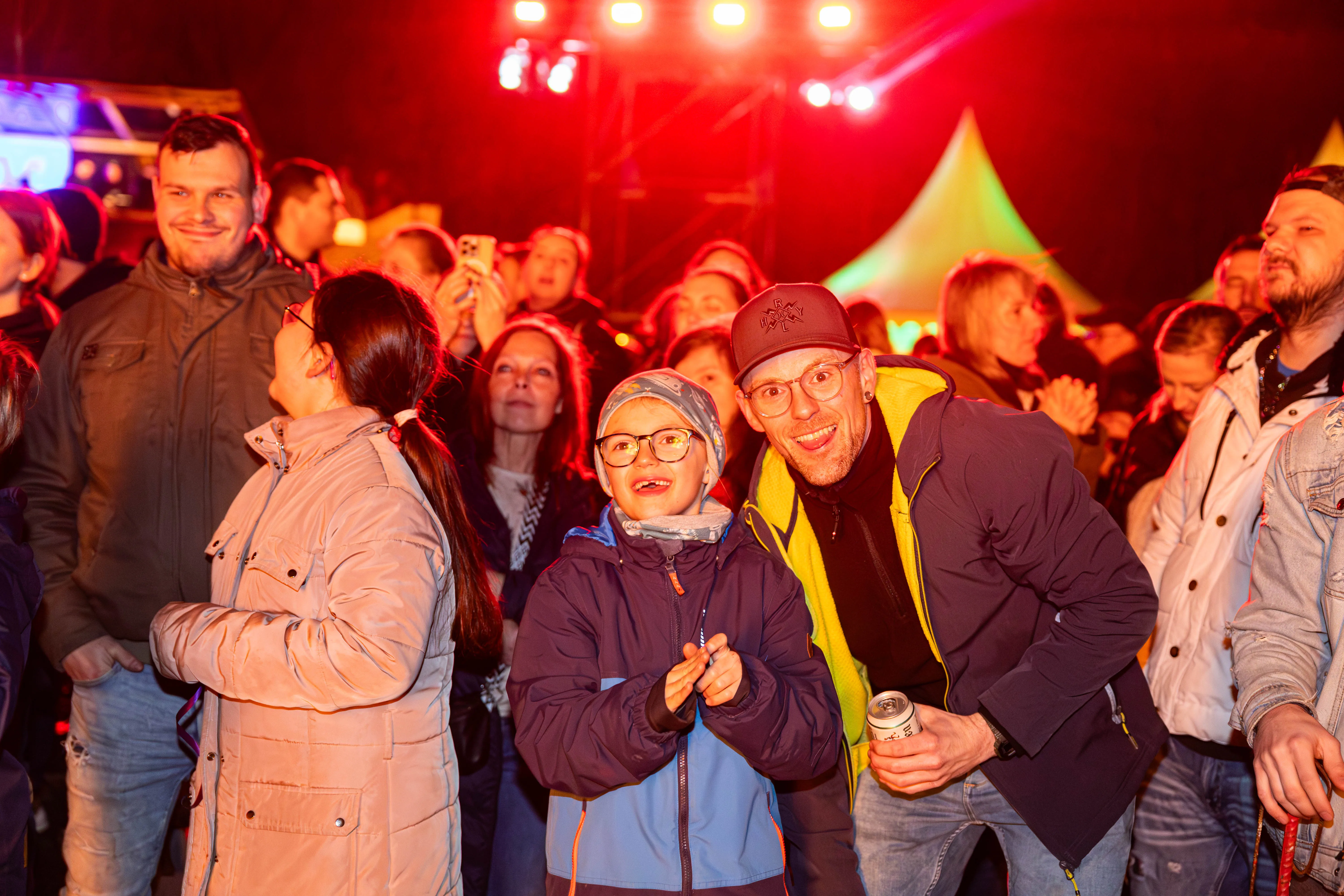 Zwei Männer und ein Kind lächeln in die Kamera, umgeben von einer jubelnden Menschenmenge vor einer Bühne mit rotem Licht. Im Hintergrund sind weitere Besucher und Zelte zu sehen.