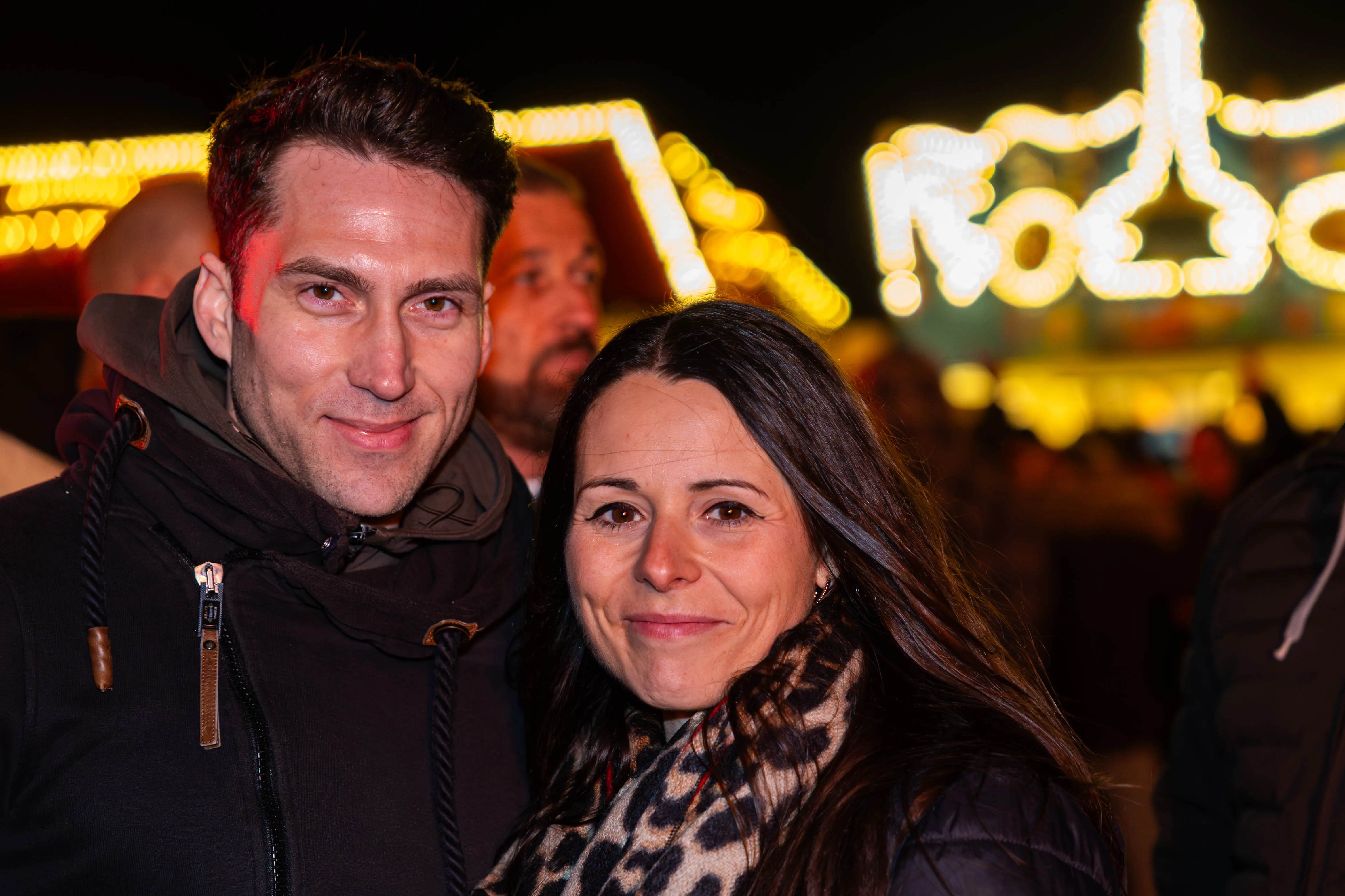 Zwei lächelnde Menschen vor verschwommenem, hell beleuchteten Hintergrund auf dem Osterfeuer-Event „Burn the Fox“ in Lüdenscheid.