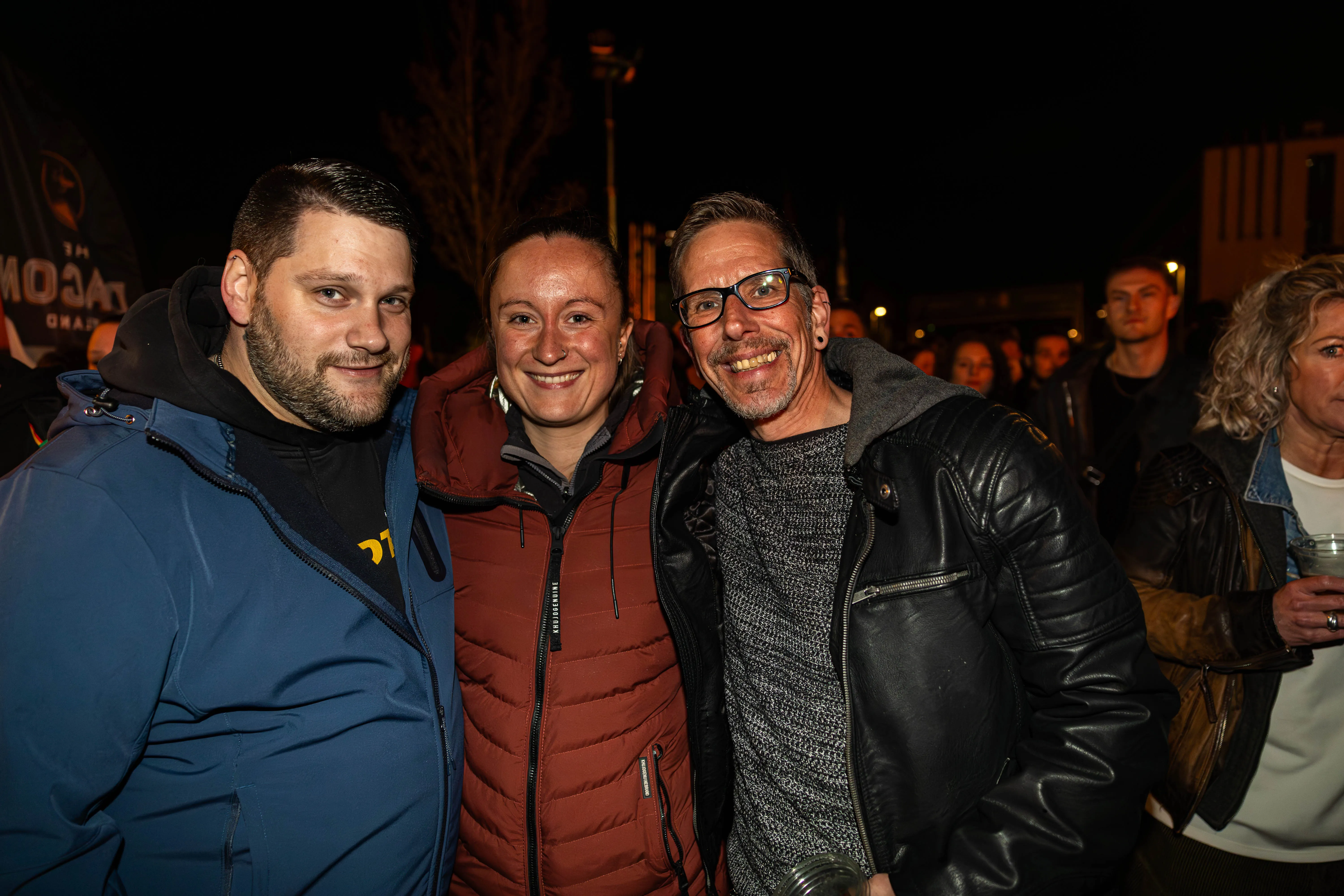 Drei lächelnde Menschen posieren eng nebeneinander für ein Foto vor einer Menschenmenge bei einem nächtlichen Osterfeuer.