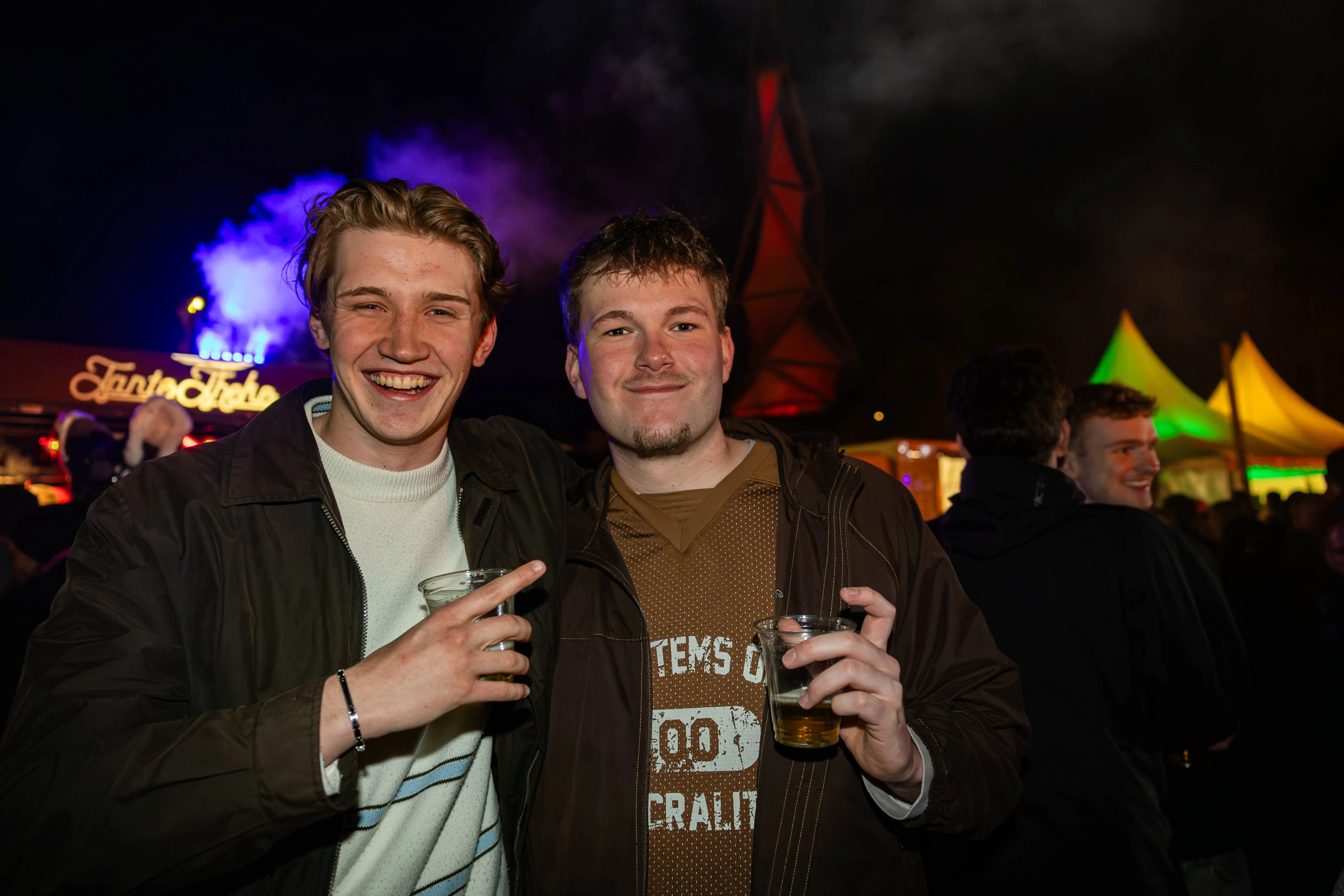 Zwei junge Männer lächeln in die Kamera und halten jeweils ein Glas in der Hand, im Hintergrund ein belebtes Osterfeuer mit weiteren Menschen und Zelten.
