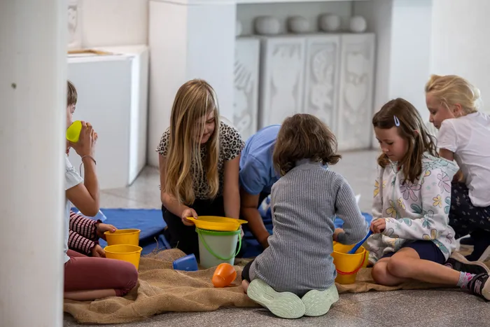 Kinder spielen auf einer Matte mit bunten Plastikeimern während einer Theateraufführung.