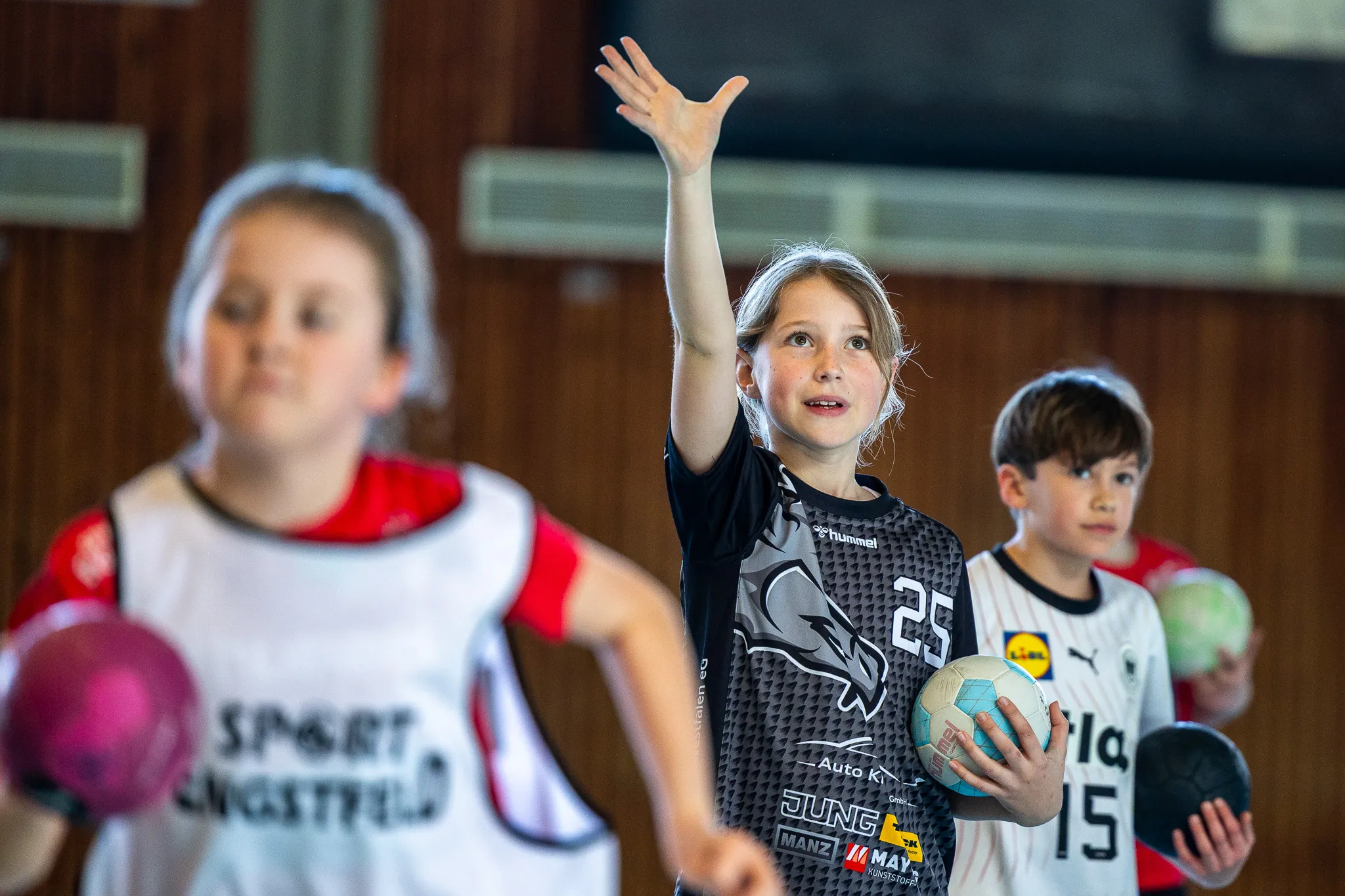 Ein Mädchen mit hochgehobener Hand und Blick auf einen Handball, umgeben von anderen Kindern in Sporthalle während eines Handballcamps.