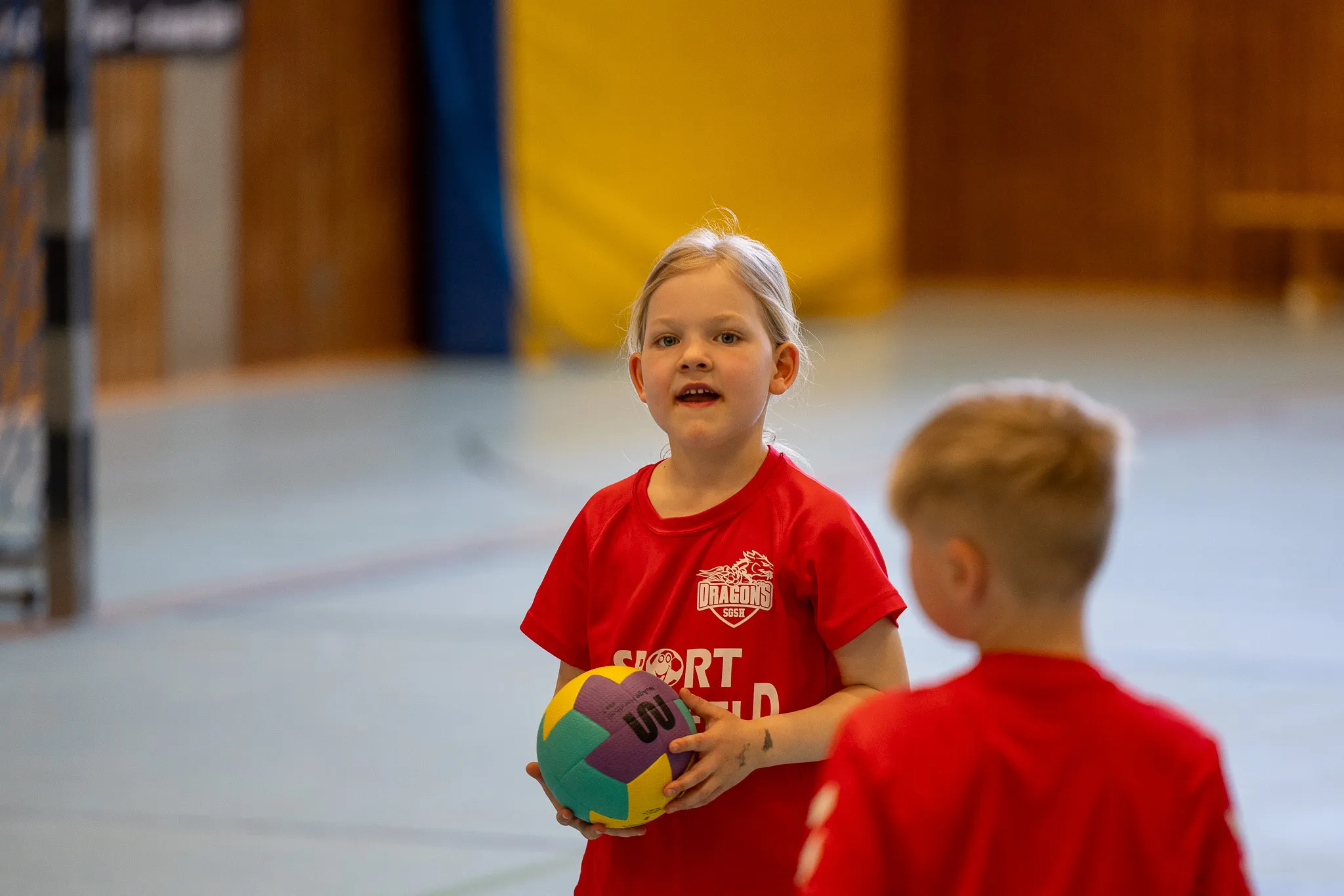 Ein Mädchen im roten Handballtrikot hält einen bunten Ball und blickt erwartungsvoll auf einen Jungen, der ihr gegenübersteht. Im Hintergrund eine Turnhalle mit Holzwand.