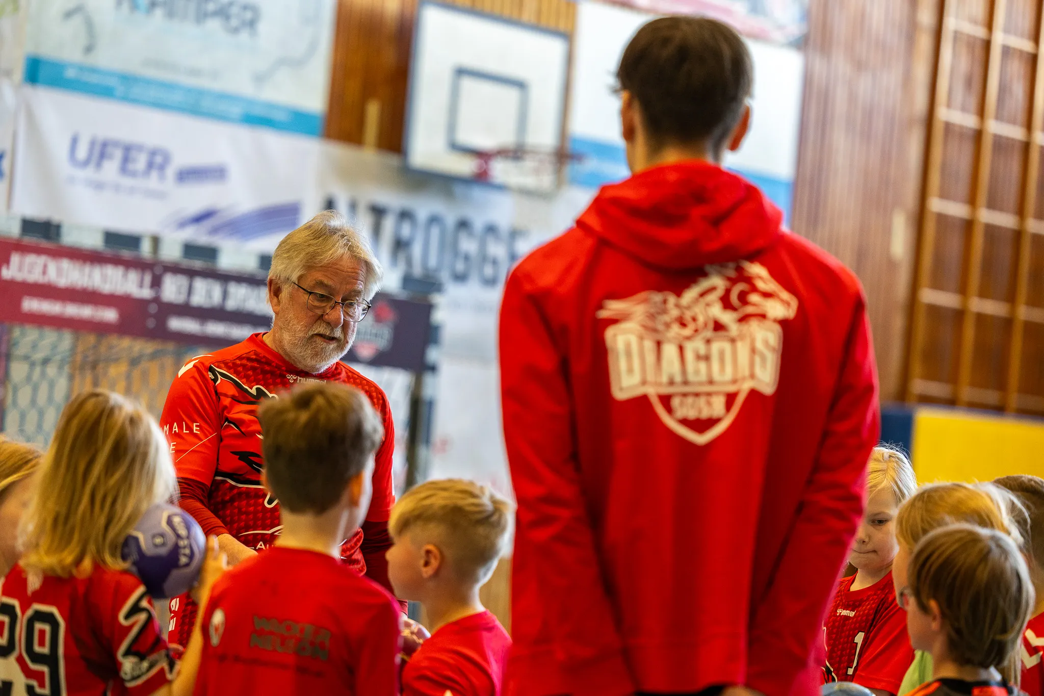 Ein Trainer spricht eine Gruppe von Kindern in roten Handballtrikots an. Im Hintergrund ein Handballtor und Werbebanner.