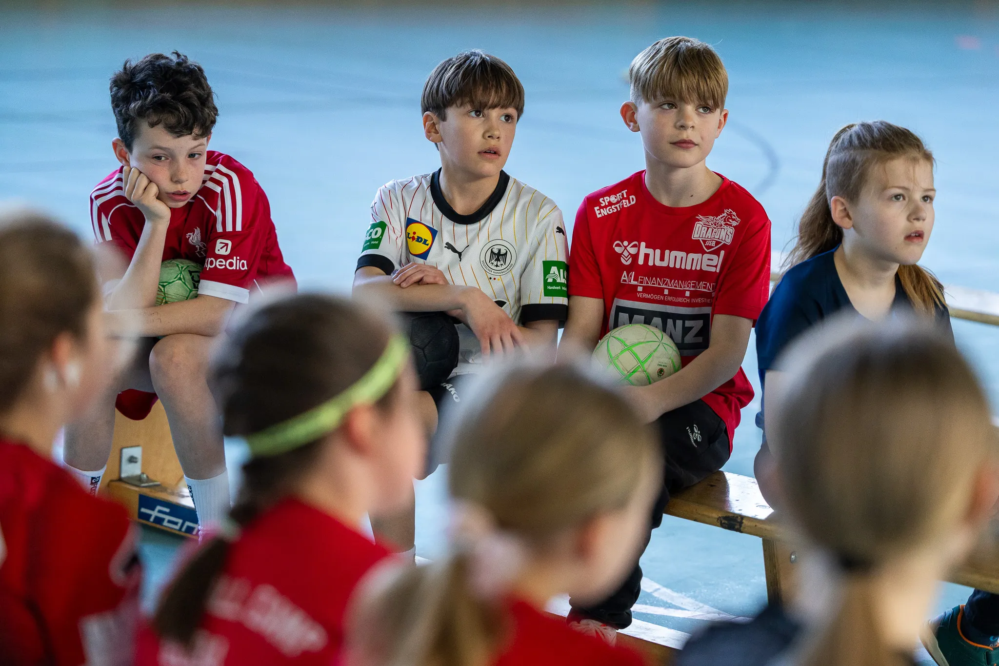 Vier Jungen sitzen auf einer Bank in einer Sporthalle und halten Handbälle. Um sie herum sitzen weitere Kinder in roten Trikots.