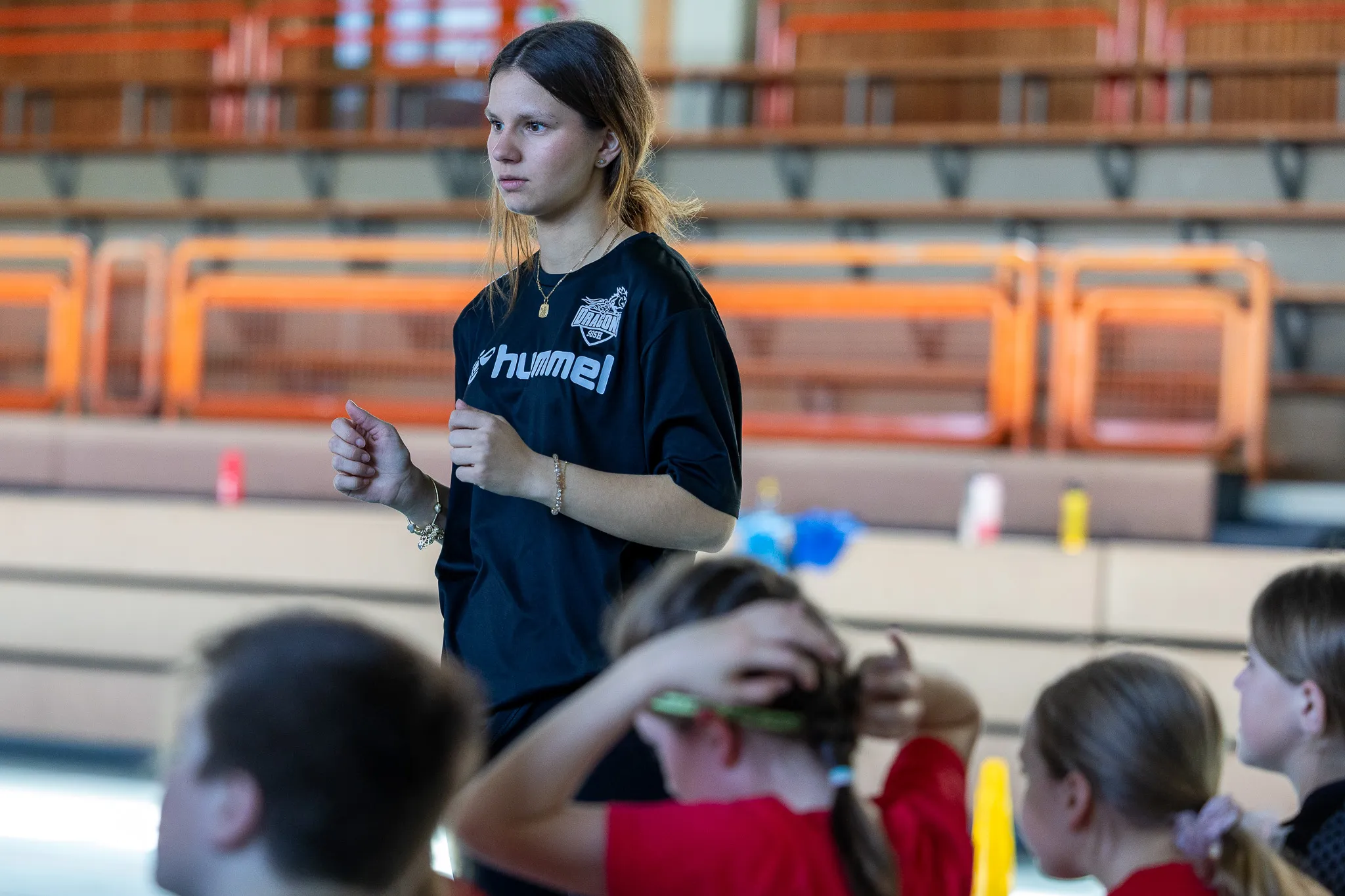 Eine junge Trainerin mit hochgesteckten Haaren steht inmitten einer Gruppe von Kindern in einer Sporthalle. Sie trägt ein Trainingsshirt mit dem Logo von Hummel und dem Schriftzug Dragons.