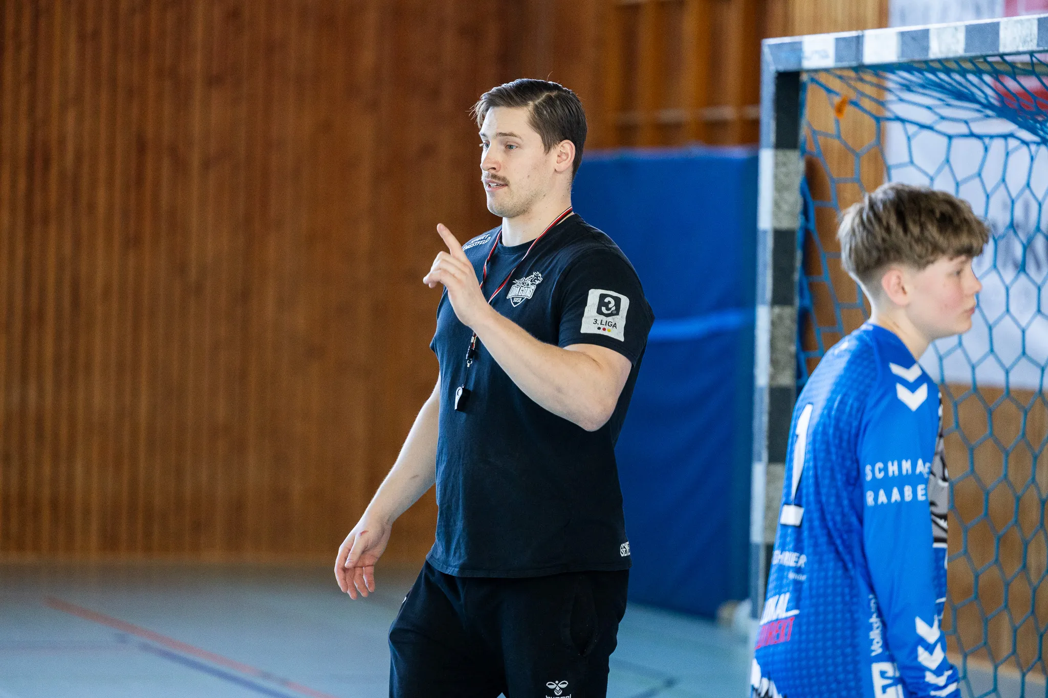 Trainer gestikuliert während eines Handballcamps in einer Sporthalle. Im Hintergrund ein Jugendspieler im blauen Trikot.